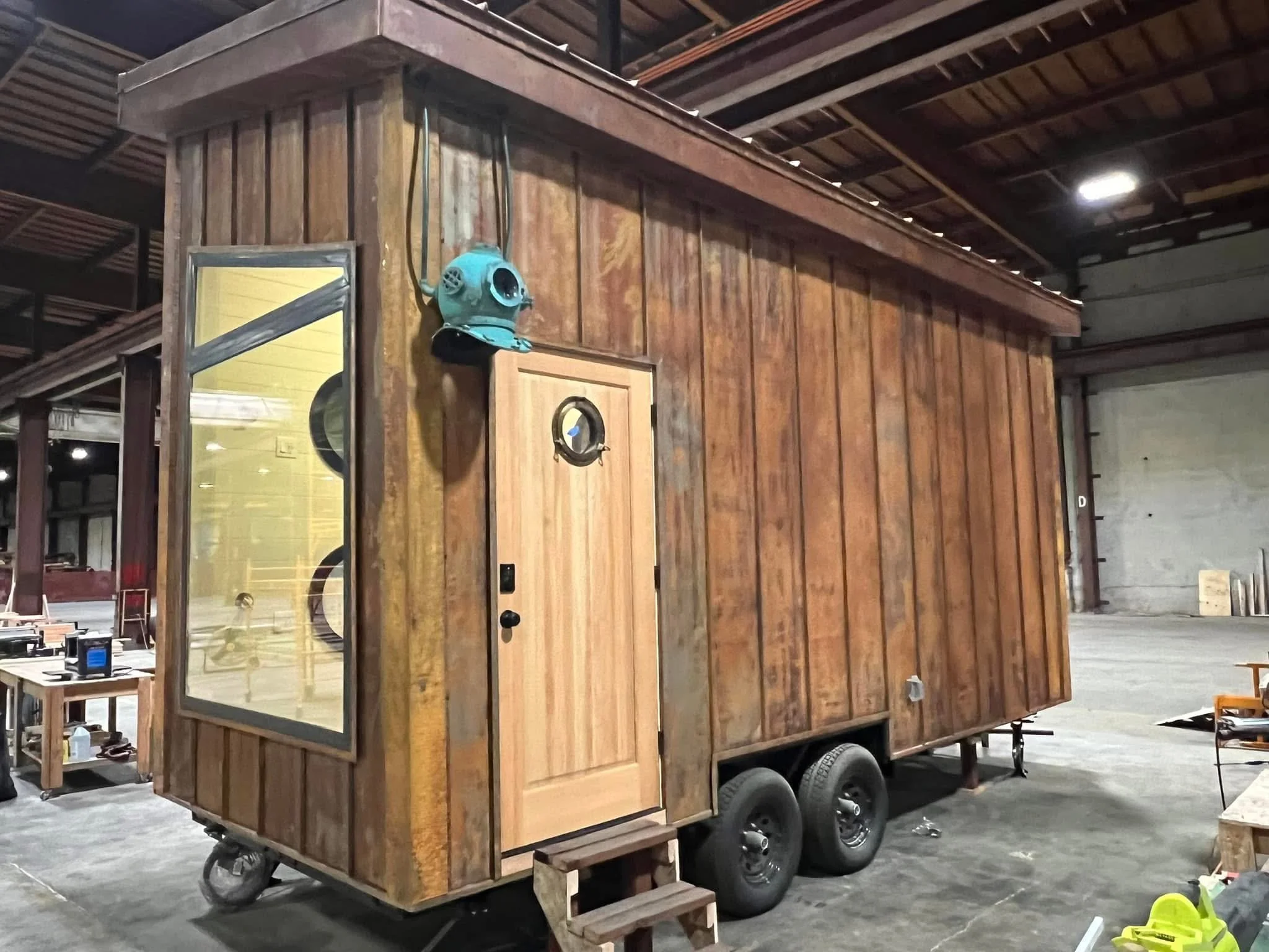 A small wooden building on wheels, resembling a tiny house or mobile cabin, inside a warehouse or industrial space.