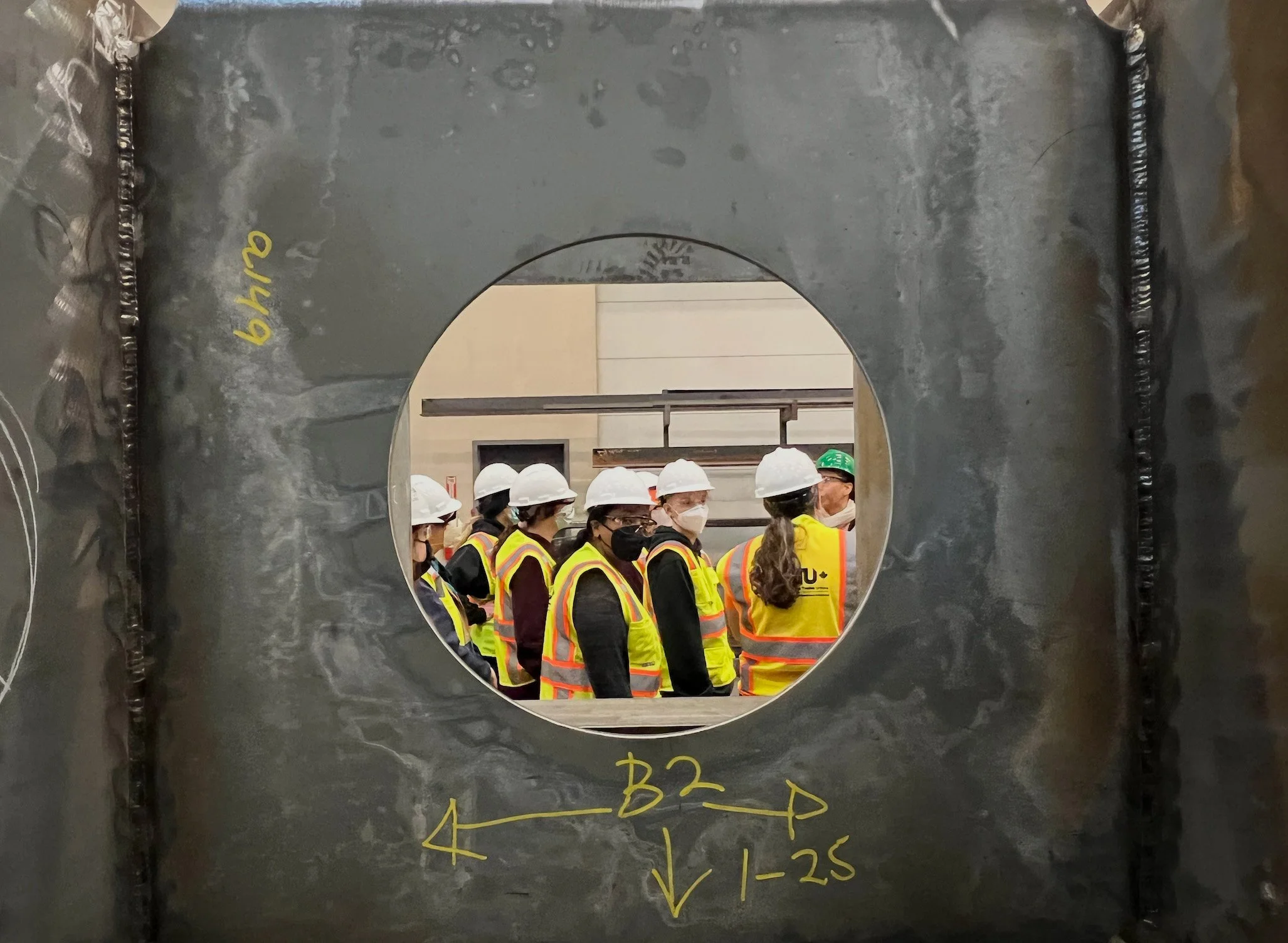 A group of construction workers wearing protective helmets and safety vests standing together, viewed through a circular hole in a black steel wall.