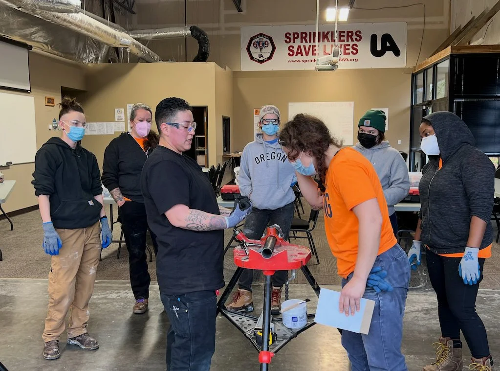Group of people wearing masks and gloves gathered around a workbench with a pipe, in a workshop or training room.