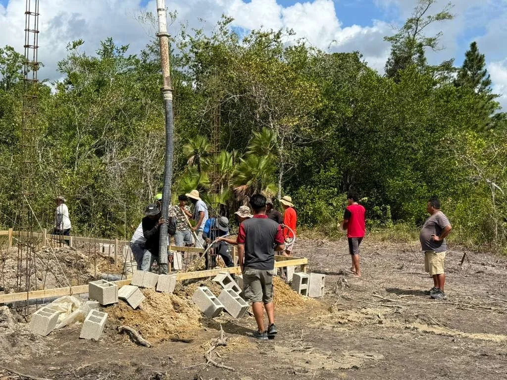 Group of people working together outdoors on a construction project with concrete blocks and wooden forms, surrounded by trees and clear sky.
