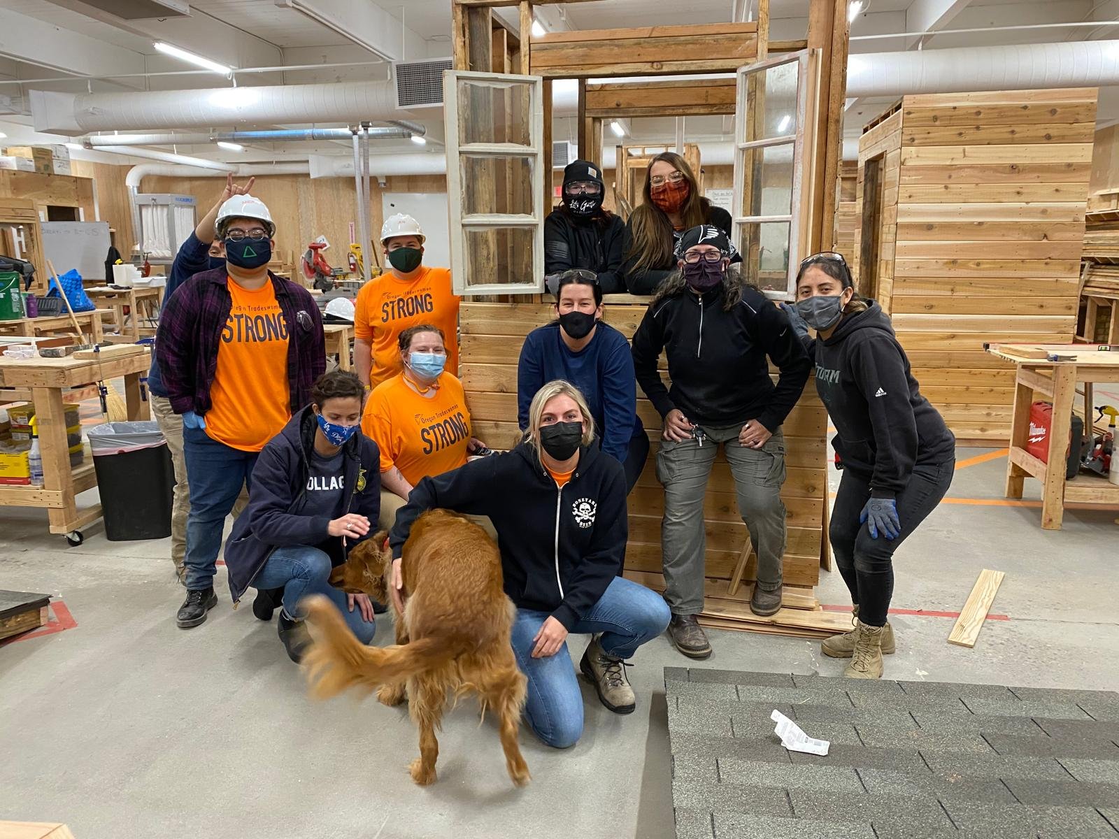 Group of people in a woodworking shop posing for a photo, some wearing safety gear and masks, with a golden retriever dog in the foreground.