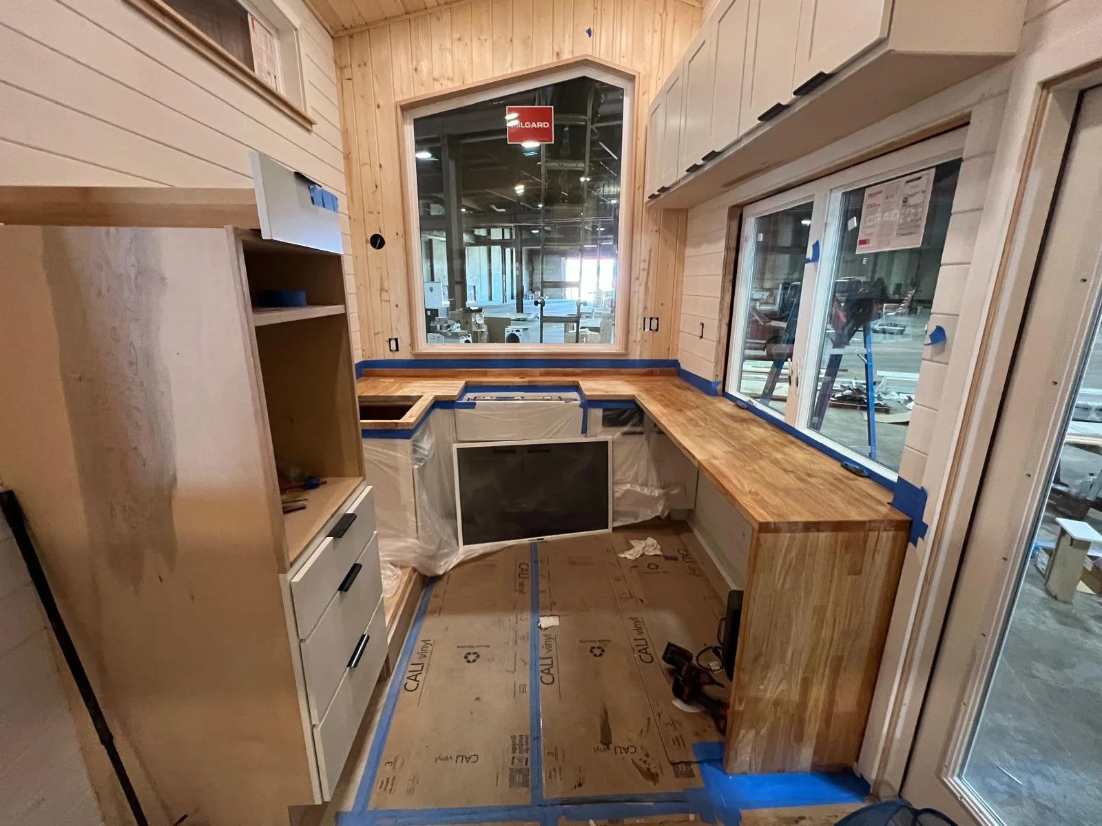 Interior of a kitchen under construction with wooden countertops, large windows, and cabinetry, with protective paper on the floor.