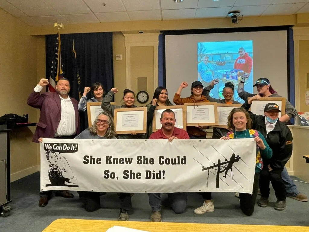 A group of people in a room holding a large banner that says "She Knew She Could So, She Did!" with a Rosie the Riveter graphic and a power line worker graphic. They are celebrating with some raising their fists, and some holding framed certificates.