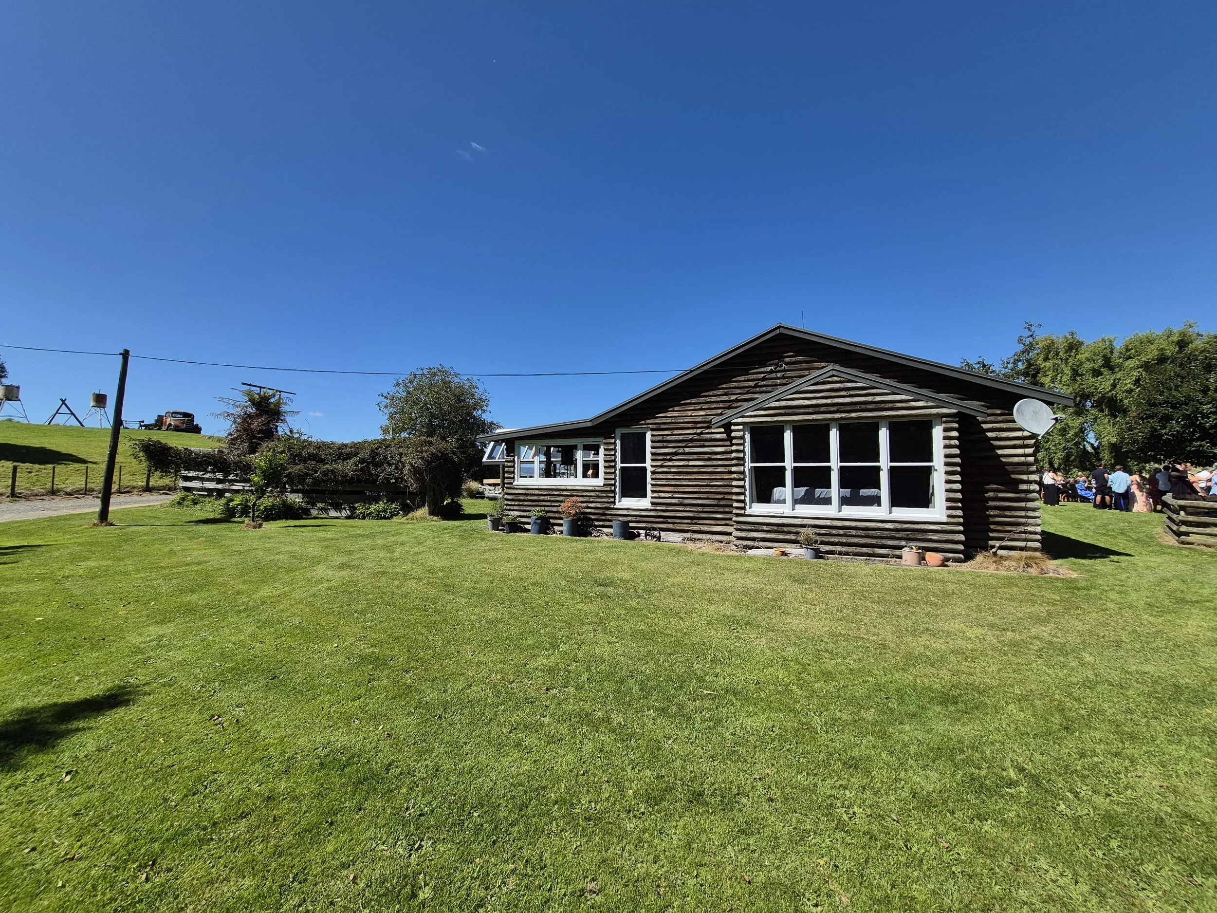 A rustic log cabin with large front windows, situated on a well-maintained grassy lawn under a clear blue sky, with a group of people gathering in the background.
