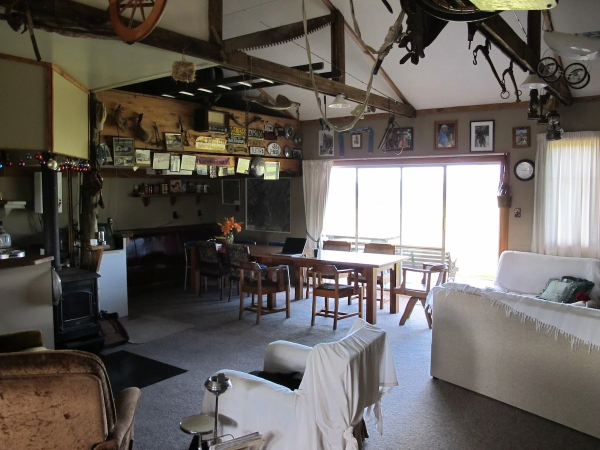 Living room with wooden ceiling beams, a dining table with chairs, a white sofa, vintage decorations, framed photos on the wall, and large windows with curtains.