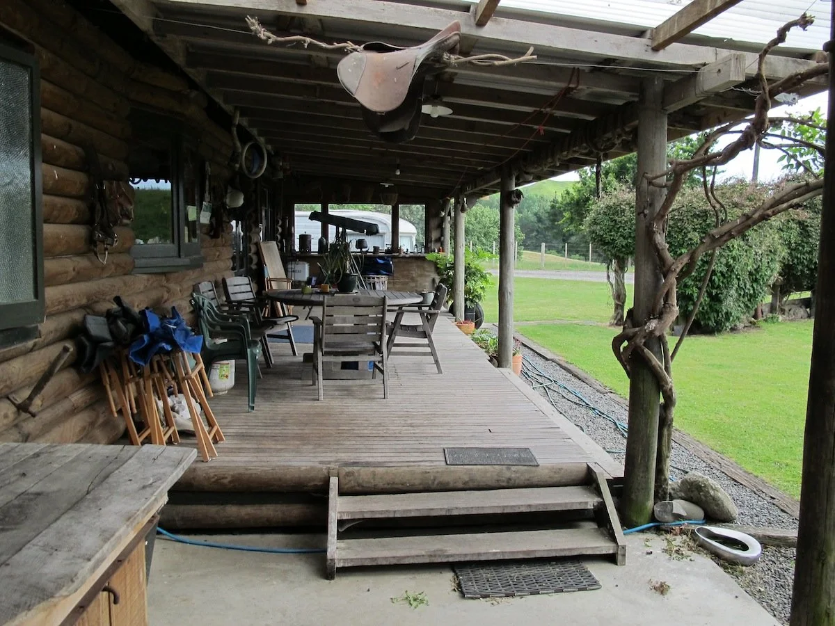 Covered wooden porch with outdoor furniture, including a table and chairs, and various gardening and outdoor tools, next to a log cabin and a grassy yard.
