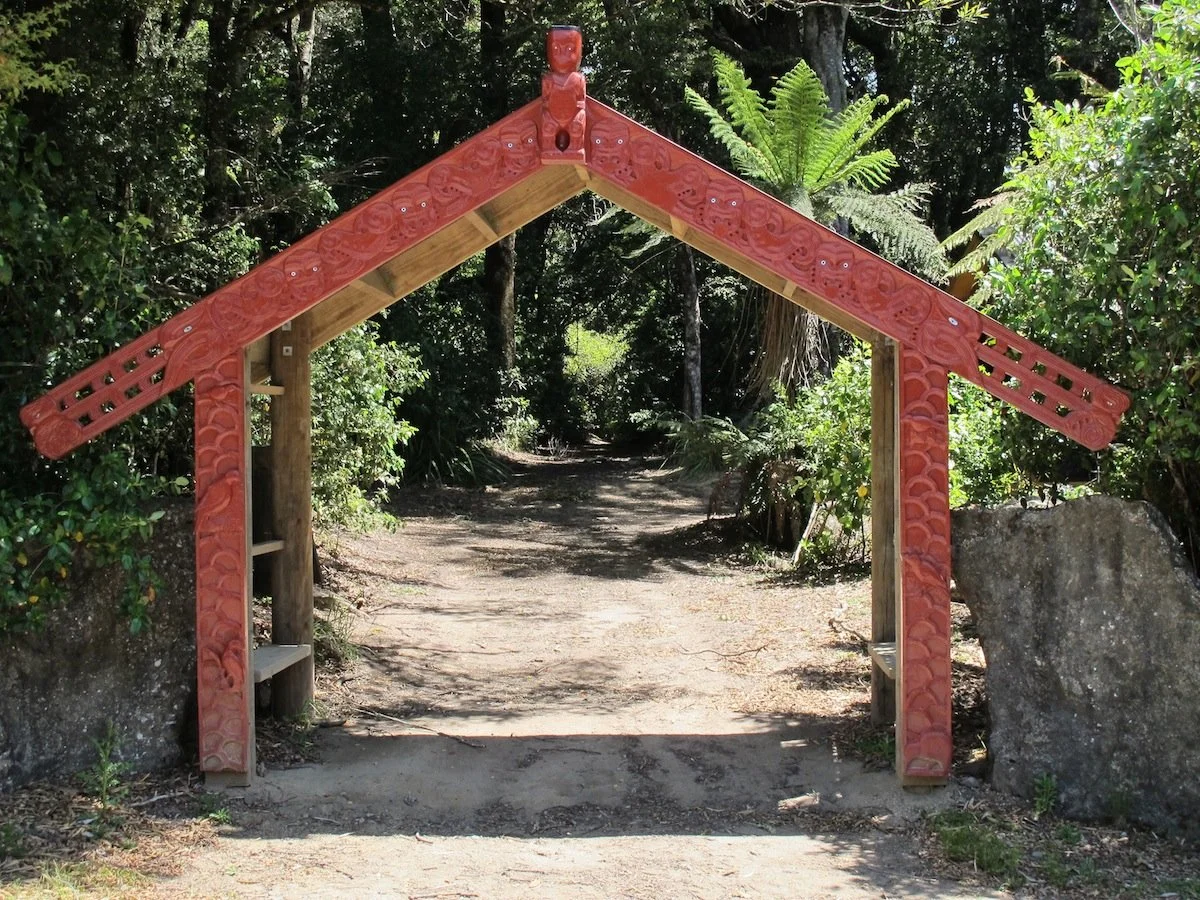 Traditional Maori carved wooden gateway at the entrance to a forest trail, with lush green trees and plants surrounding it.