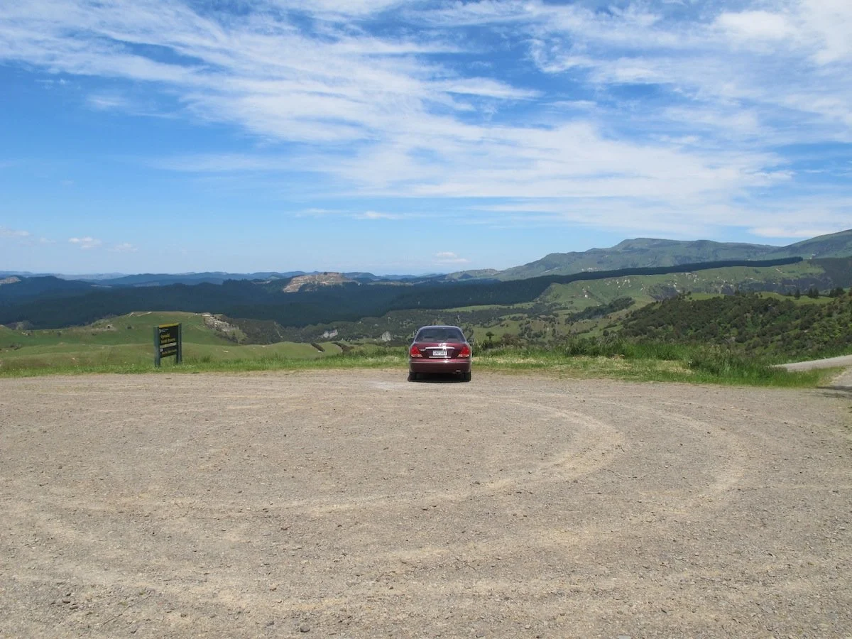 A single red car parked on a dirt lot overlooking green rolling hills and mountains under a partly cloudy blue sky.