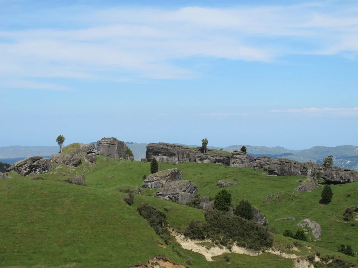 Green hilly landscape with large rocks and sparse trees under a blue sky with some clouds.