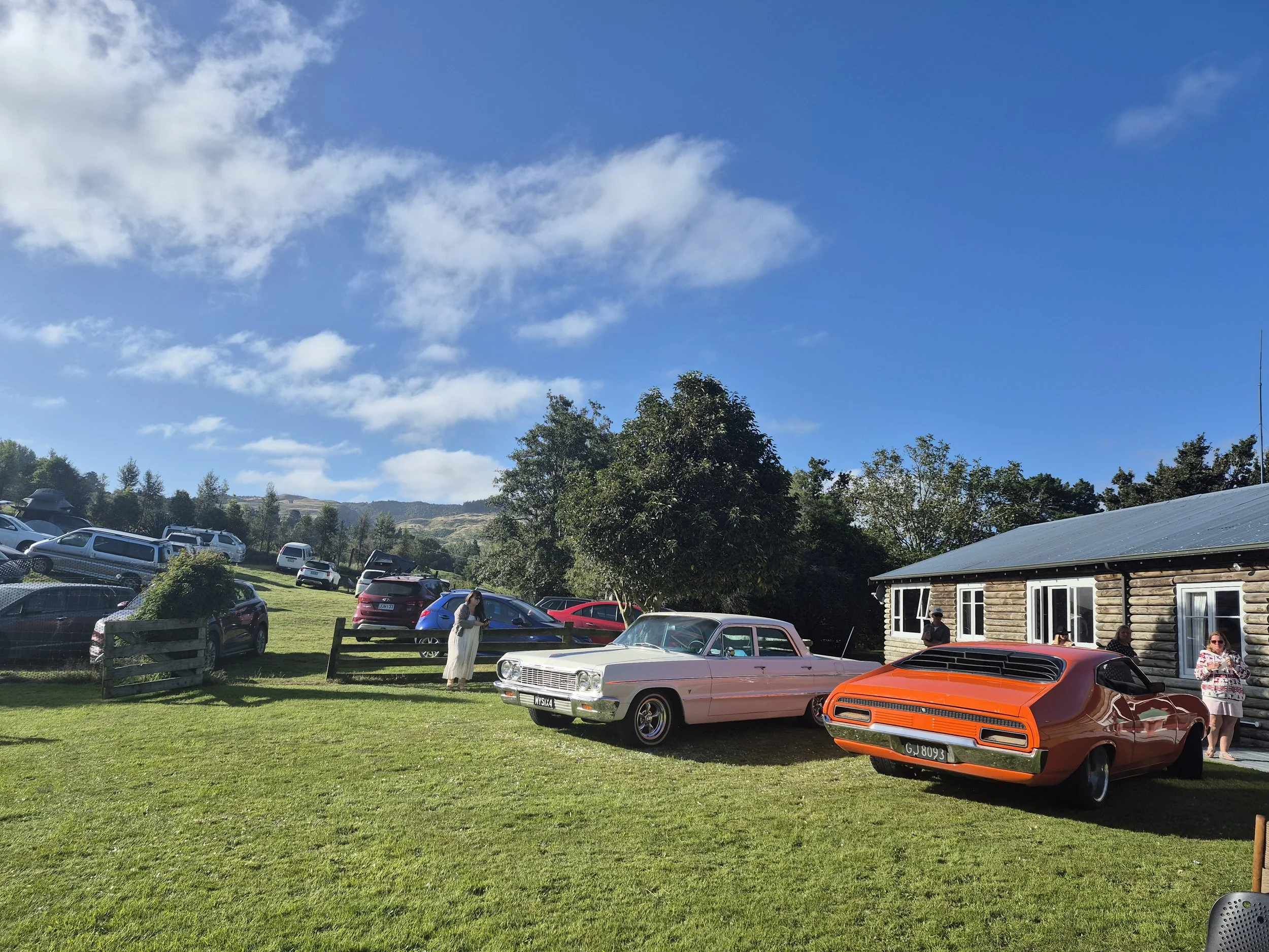 A scene of a grassy area with vintage cars and several people, next to a wooden building under a blue sky with some clouds. There are parked cars and trees in the background.