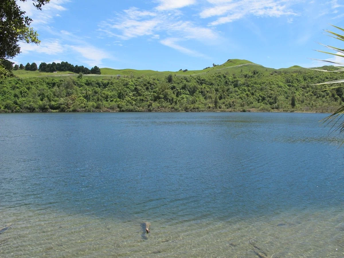 Lake Tutira with clear water in the foreground, surrounded by green hills and trees under a partly cloudy blue sky.