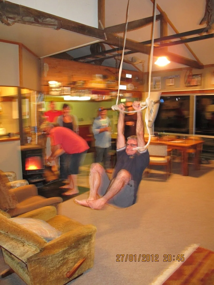 A man is swinging on a rope hung from the ceiling inside a cozy, rustic living room. There are several people in the background, some standing and chatting, in a warm, relaxed atmosphere.