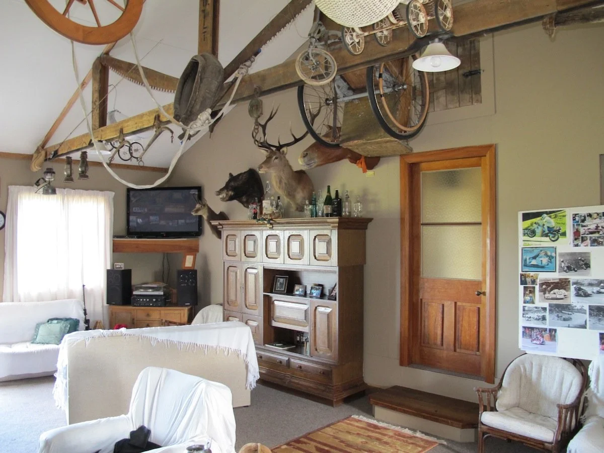 Living room with a mounted TV, wooden cabinet, white armchair, and various animal heads on the wall. Canoe and bicycle hanging from the ceiling. Photographs and decorations on the walls.
