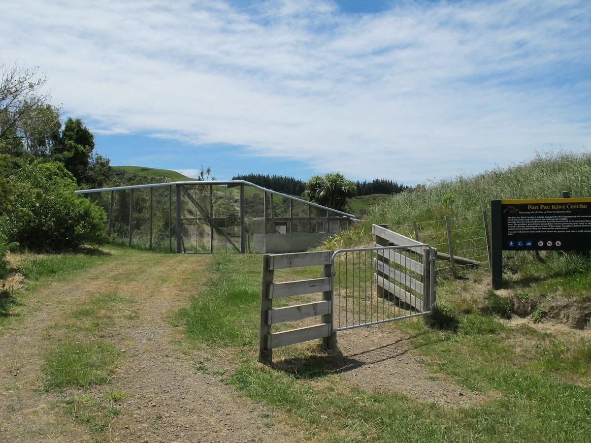 Entrance to the Pan Pac Kiwi Croche sanctuary with a small gate, sign, and green grassy landscape under a partly cloudy sky.