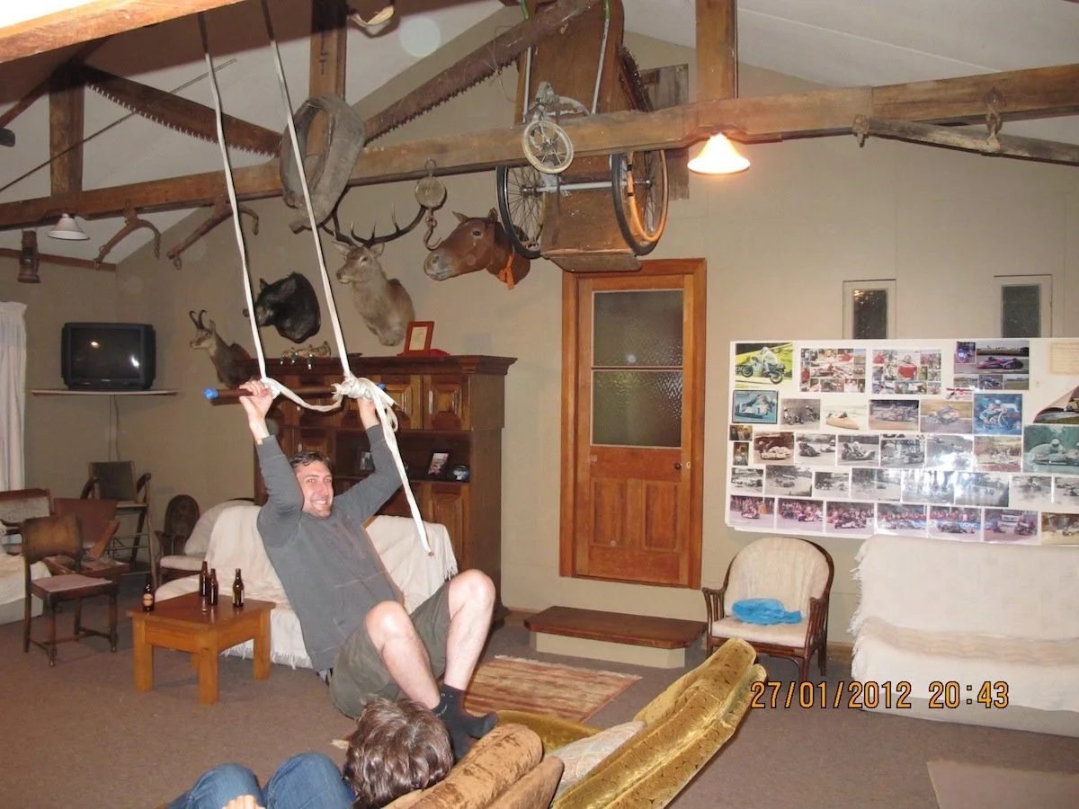 A man sitting on a couch holding onto a rope swing inside a rustic living room with animal mounts on the wall and a collage of photos on a whiteboard.