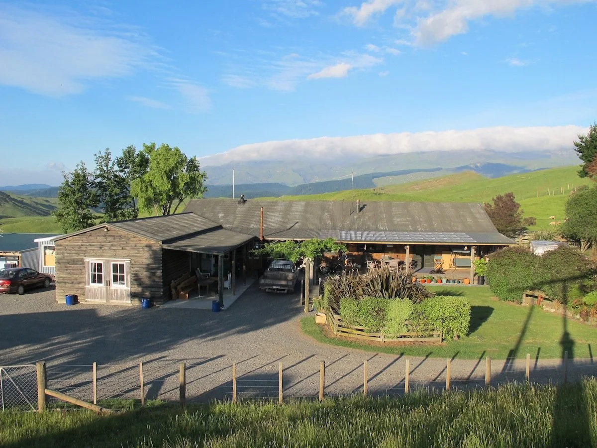 A rustic barn with a gravel driveway, surrounded by green grass, bushes, and trees, with rolling hills and mountains in the background under a partly cloudy sky.