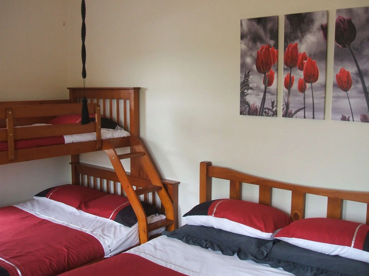 Bedroom with two beds, one on the floor and a bunk bed, both with red, black, and white bedding. There is a three-panel canvas wall art featuring red tulips on a cloudy sky background.