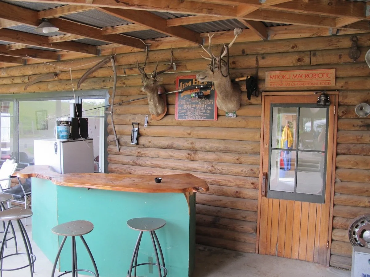 Interior of a rustic bar or restaurant with log walls, featuring two mounted deer heads with antlers, a wooden door with glass, a counter with stools, and various signs and decorations on the wall.