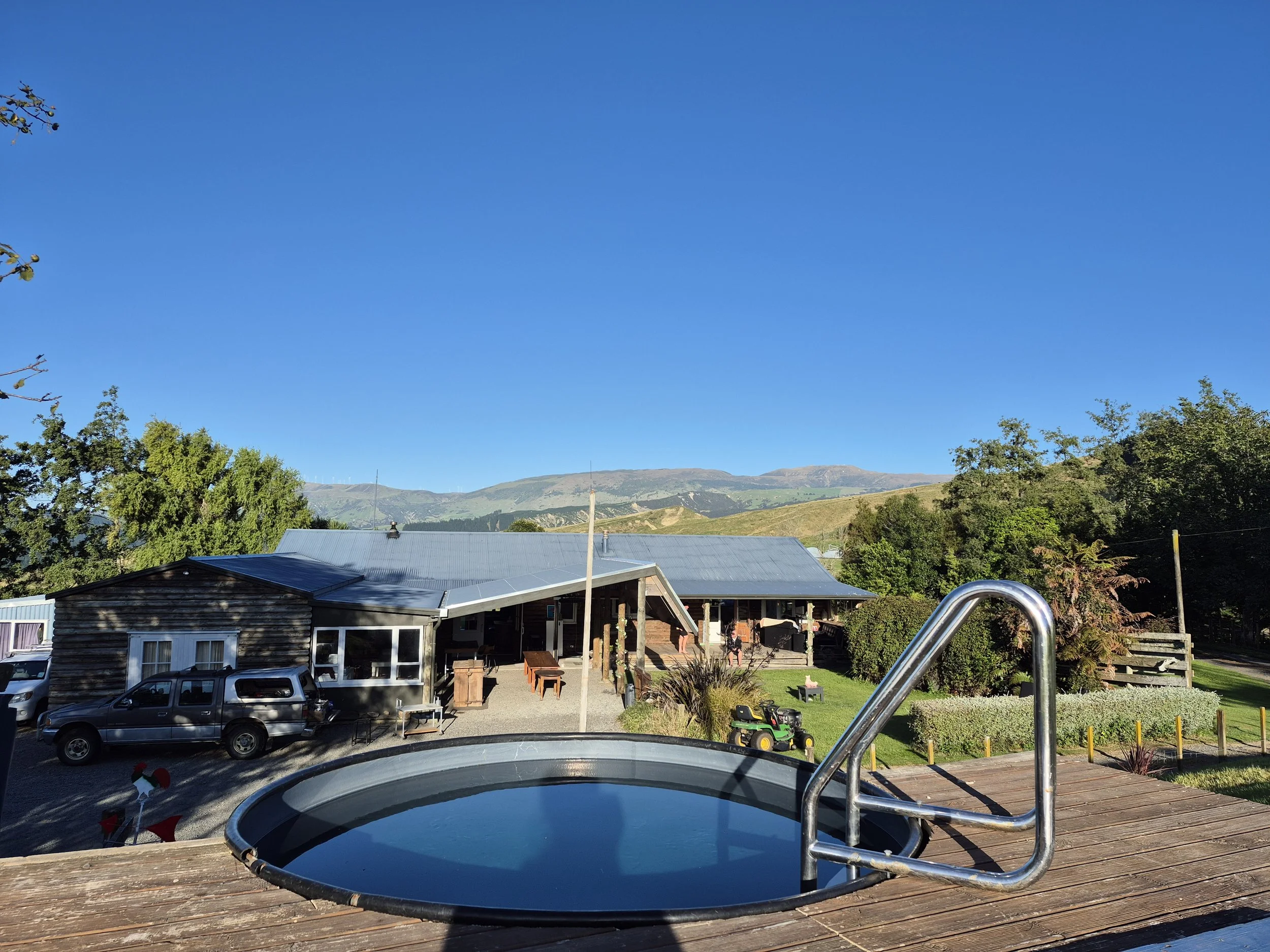 View from a deck with a small pool and metal handrail overlooking a rural area with a building, trees, vehicles, and mountains in the distance under a clear blue sky.