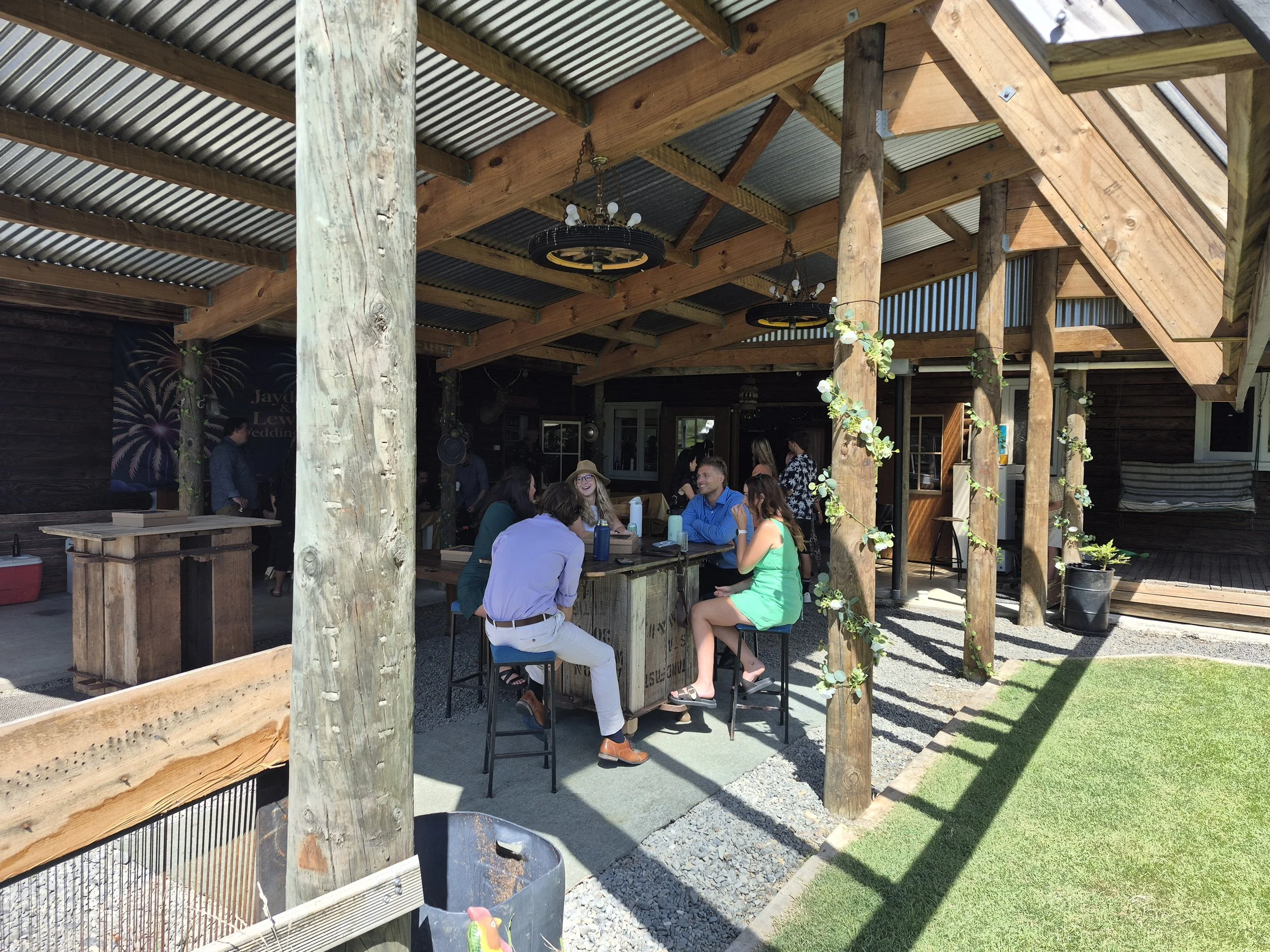 People sitting and standing around a rustic outdoor bar under a wooden and metal roof at a casual gathering or party.