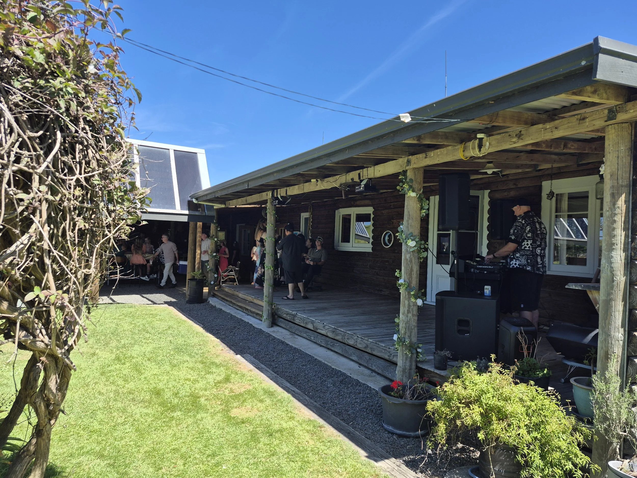 Outdoor gathering at a house with a wooden porch, people dancing and socializing, a DJ playing music, and potted plants on the porch on a sunny day.