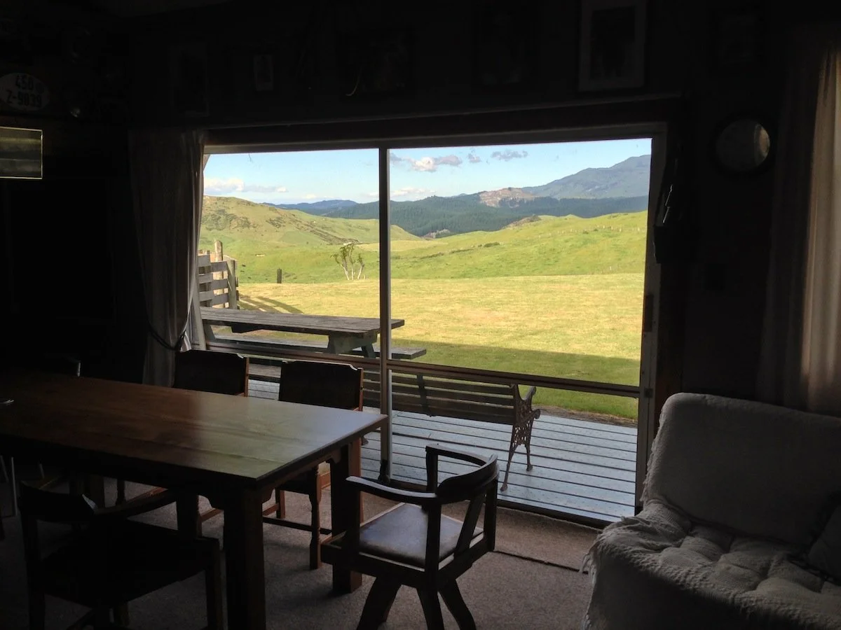 Interior of a cozy room with a wooden dining table, chairs, and a white couch, viewing a lush green landscape with rolling hills and mountains through a sliding glass door.