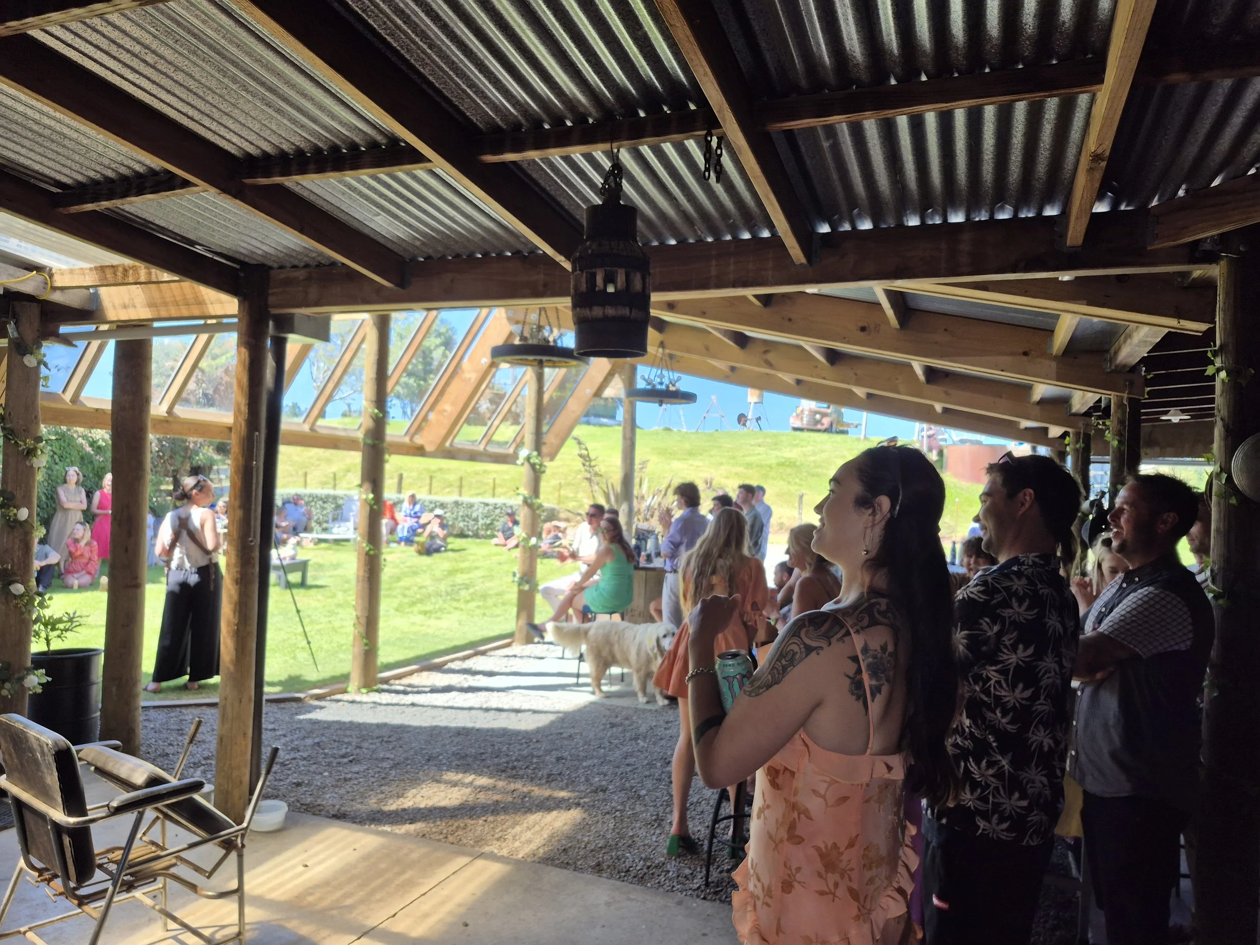People gathered at an outdoor event under a rustic wooden shelter with a view of a grassy hillside and a blue sky, some sitting and others standing, with a dog on a leash and a woman in a floral dress holding a drink.