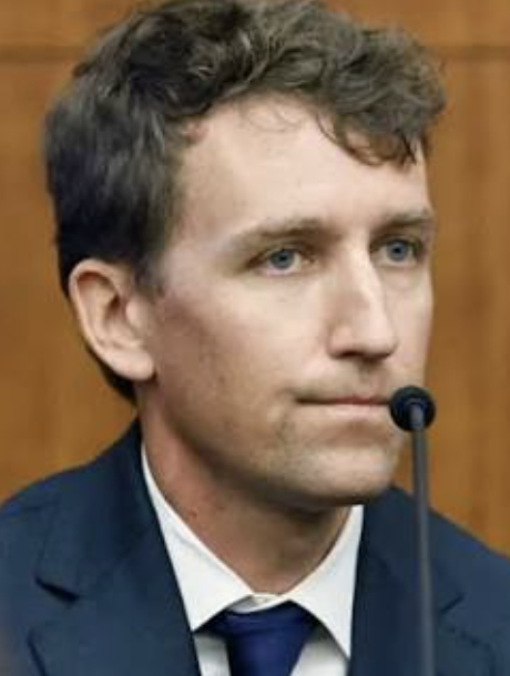 A man with light skin, short wavy brown hair, and blue eyes, wearing a dark suit, white shirt, and dark tie, sitting in front of a wooden background with a microphone near his mouth.

Patrick Clancy, July 2026