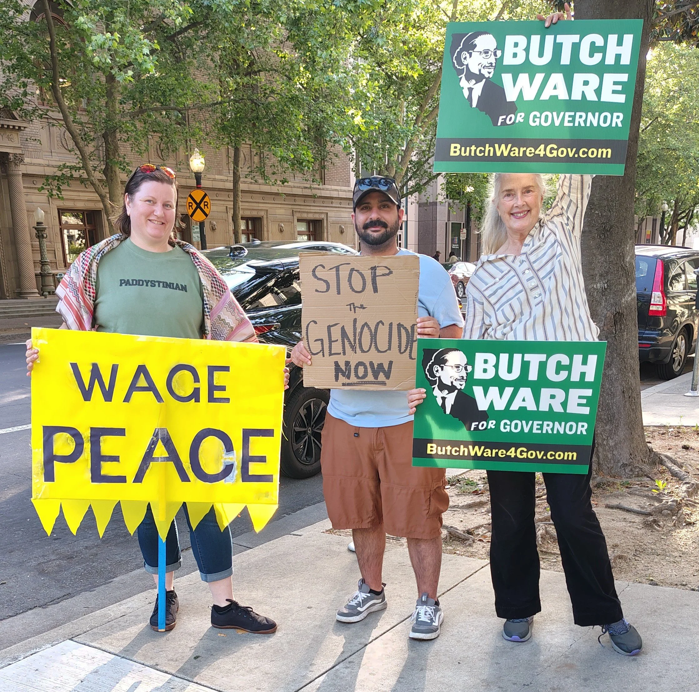 A protest with three people holding signs, including one that reads 'WAGE PEACE' in yellow letters on a yellow background. Another person holding a cardboard sign with handwritten text that says 'STOP THE GENOCIDE NOW.' A third person holds a large green sign that promotes a political campaign for Butch Ware, with his portrait and a website URL. The protesters are standing outdoors on a sidewalk with trees and city buildings in the background.