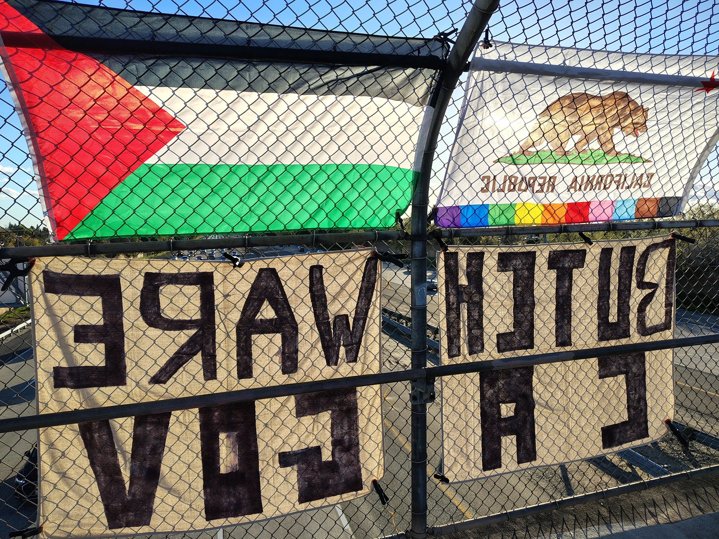 A fence with two cloth banners hanging behind it. One banner shows the Palestine flag with black, white, green, and red colors, and the other banner has a brown bear walking on green grass with the text 'CALIFORNA REPUBLIC' and rainbow-colored stripes at the bottom.