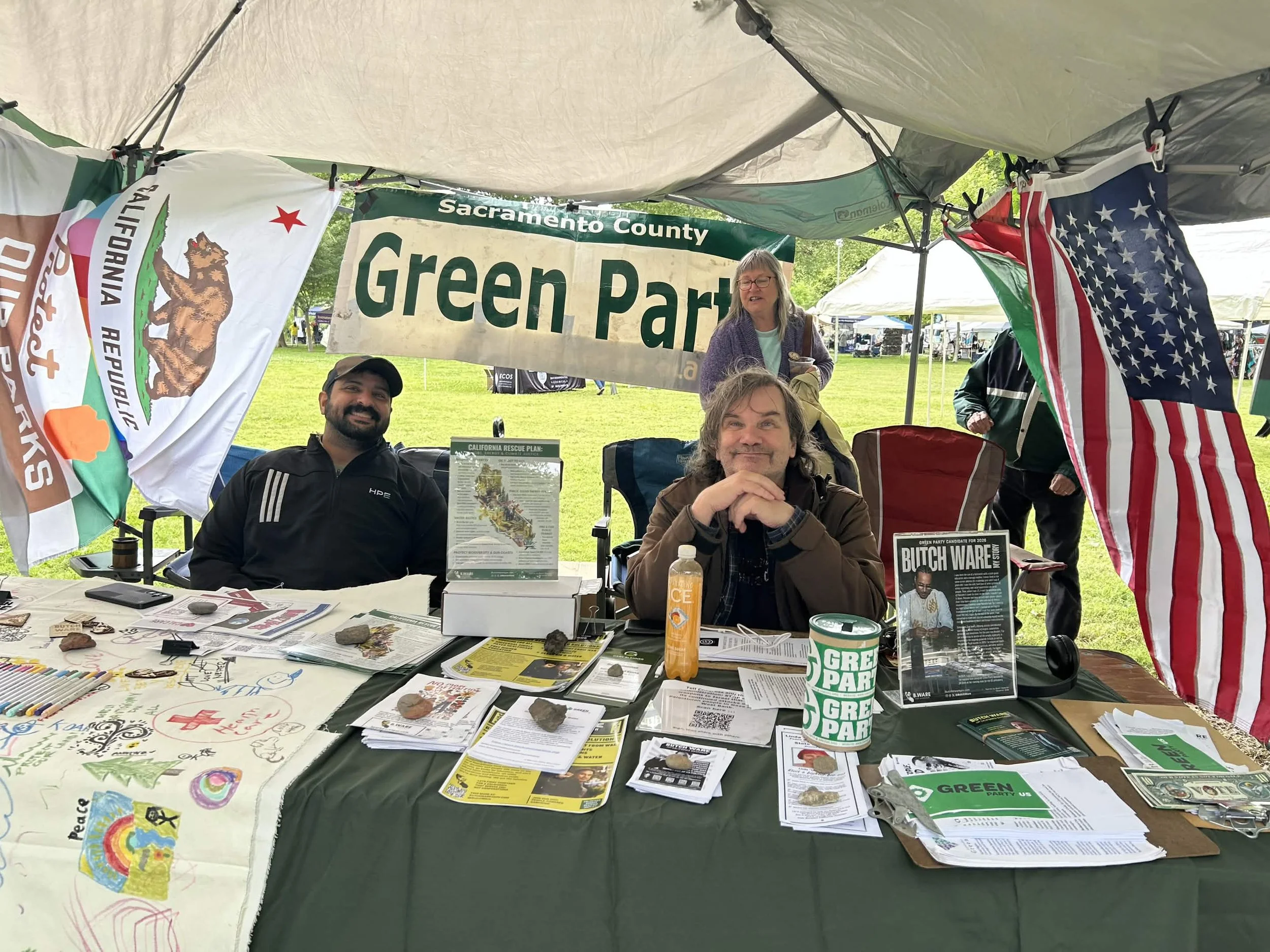 People sitting at a table under a tent at a Green Party event. The table has flyers, posters, and various informational materials about environmental issues and political candidates. Flags of California, the United States, and the Green Party are visible.