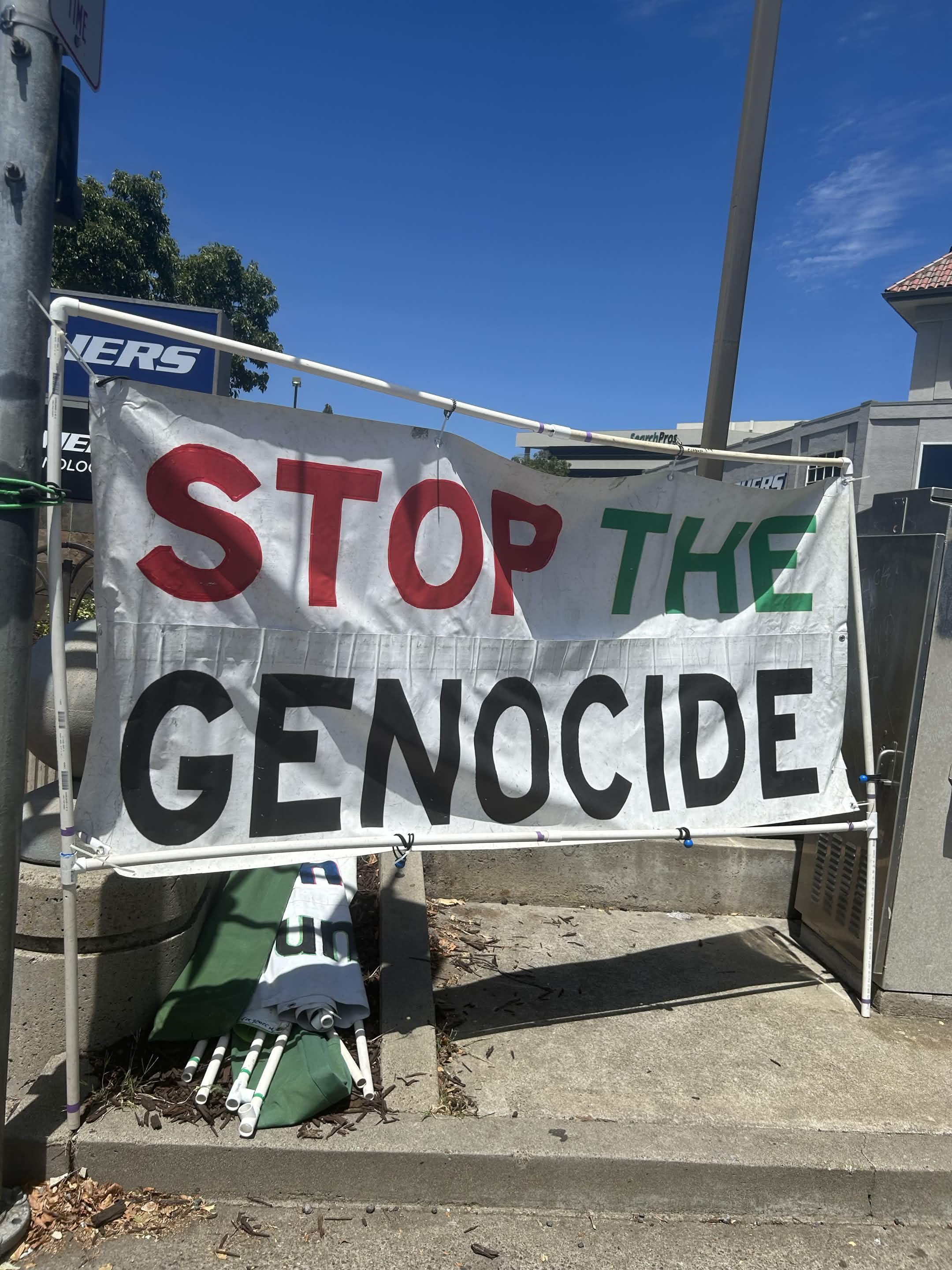 A makeshift protest sign on a metal frame that reads 'STOP THE GENOCIDE.' Several white and green poles are leaning against the frame, with some rolled-up banners and bags on the ground below. The background shows a clear blue sky and parts of nearby buildings and trees.
