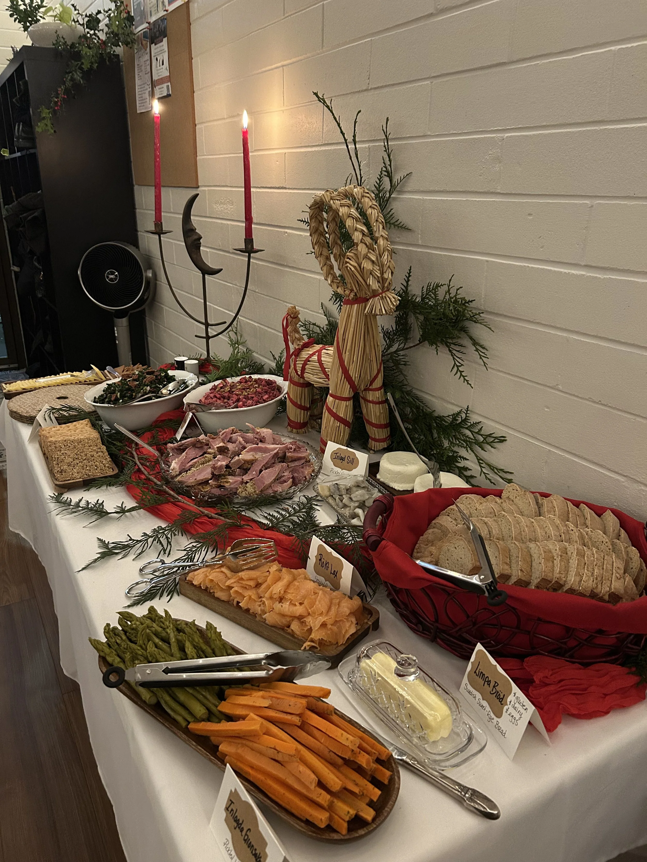 Holiday buffet table with assorted breads, smoked salmon, vegetables, sliced meats, cheeses, and festive decorations including a straw reindeer and greenery.