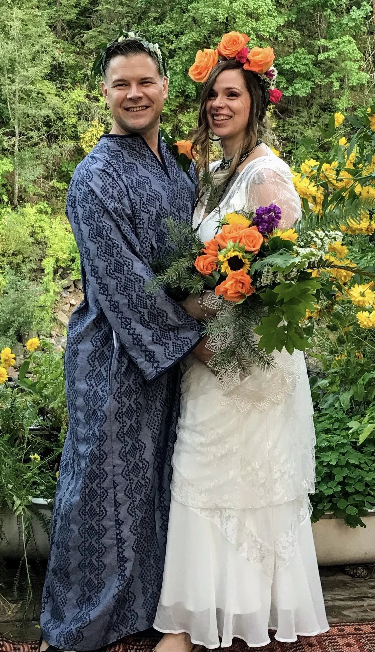 A happy couple in wedding attire, with the groom in traditional blue patterned clothing and the bride in a white lace dress, holding a colorful bouquet of flowers, standing outdoors among green trees and yellow flowers.