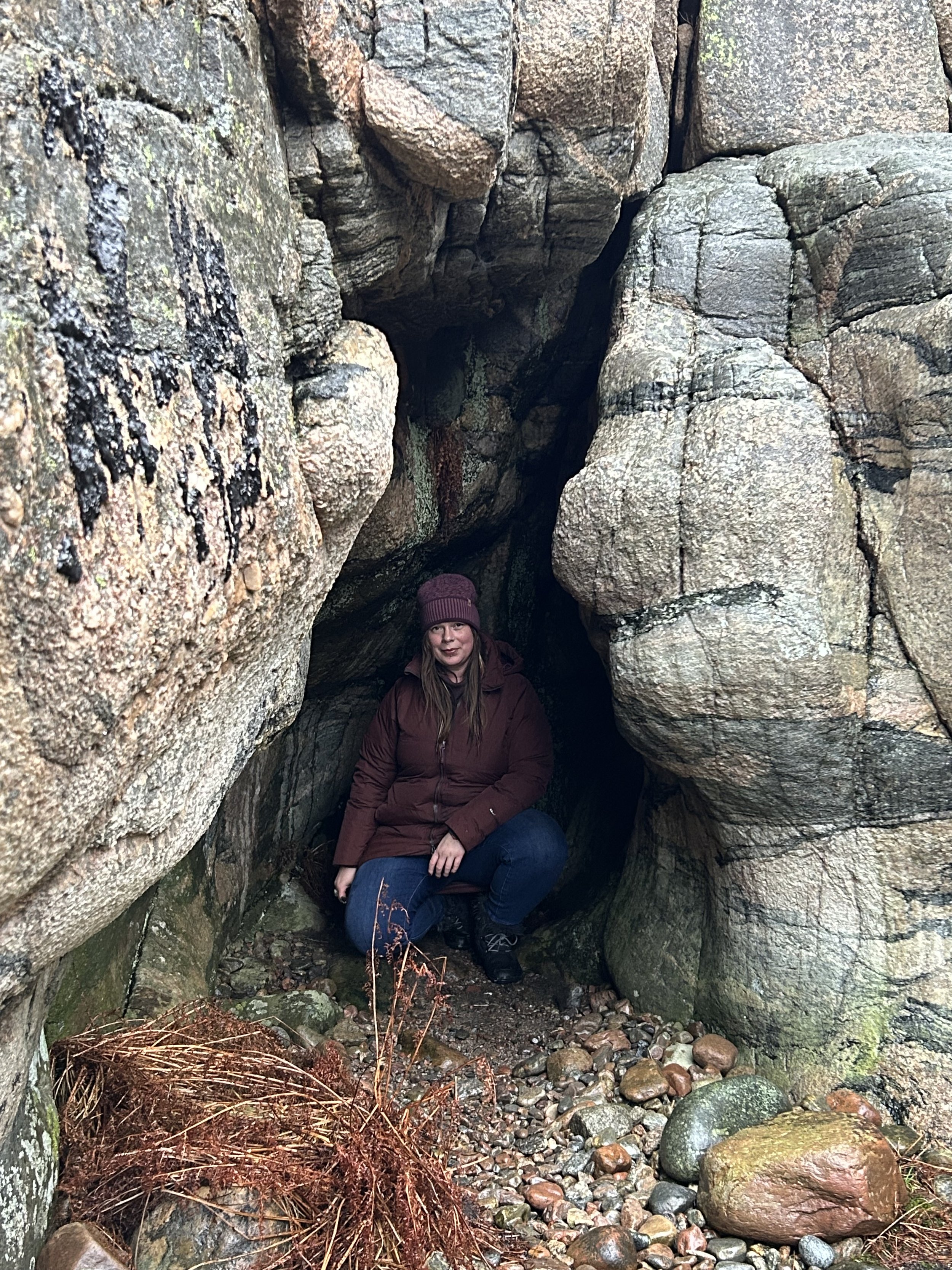 A woman crouches inside a narrow rocky crevice, surrounded by large, textured stones and pebbles, wearing a maroon jacket, jeans, and a knit hat.