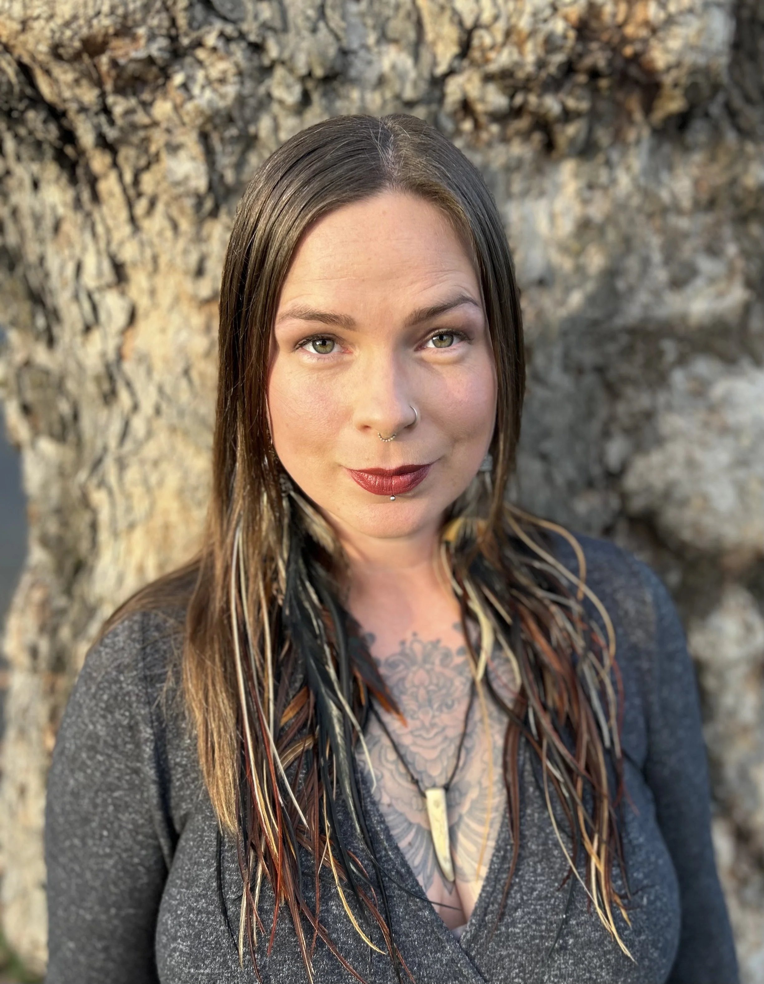 A woman with straight brown hair and light skin, standing in front of a tree trunk. She has green eyes, a nose ring, a labret piercing, and is wearing a dark gray top. She has tattoos on her chest and is wearing a necklace with a pendant. The woman is looking at the camera with a slight smile.