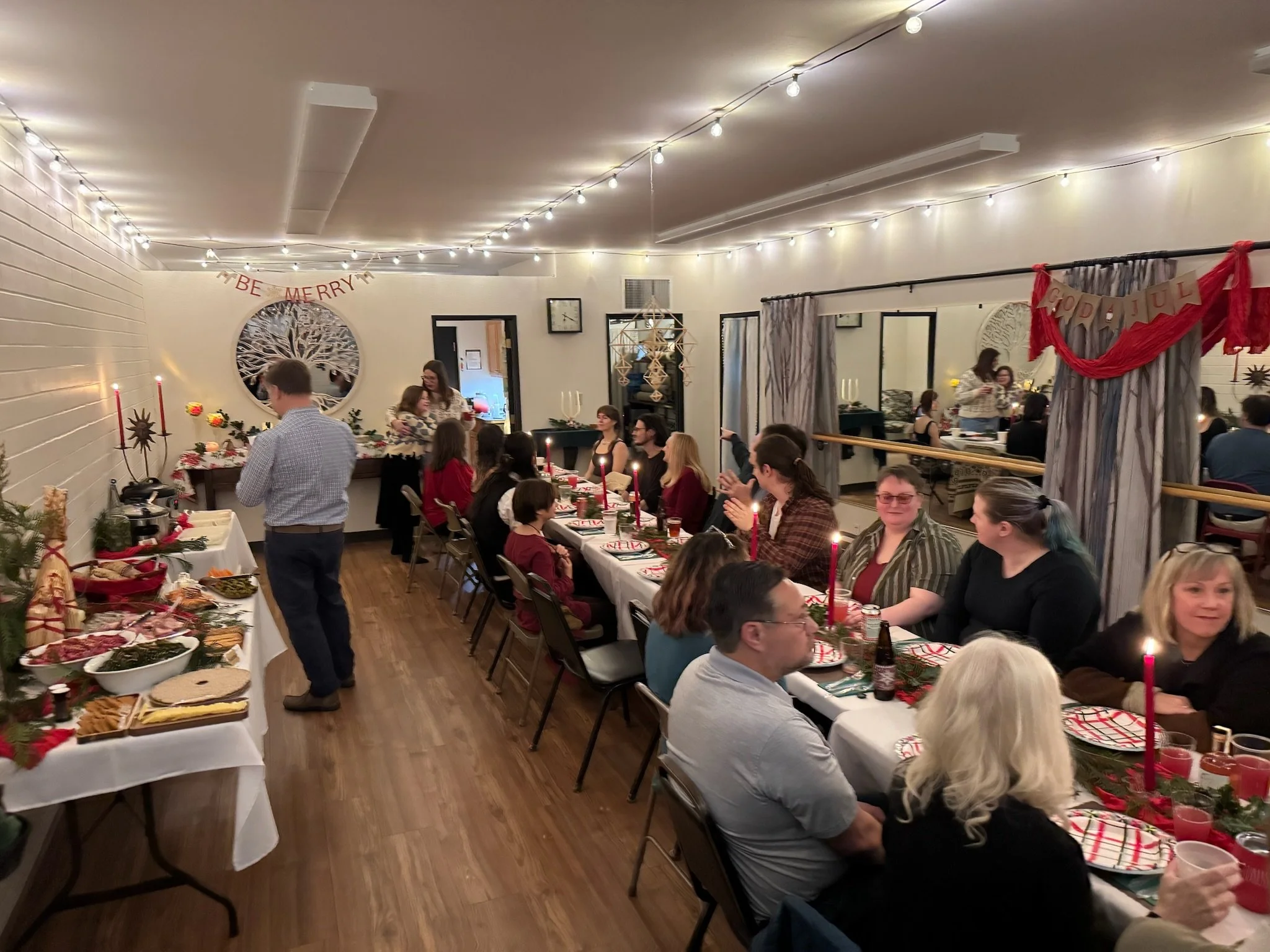 A Christmas dinner party with guests seated at a long table decorated with red candles, holiday-themed tableware, and greenery. Hanging holiday banners read 'Be Merry' and 'God Jul'. The room has string lights, curtains, and festive decorations.
