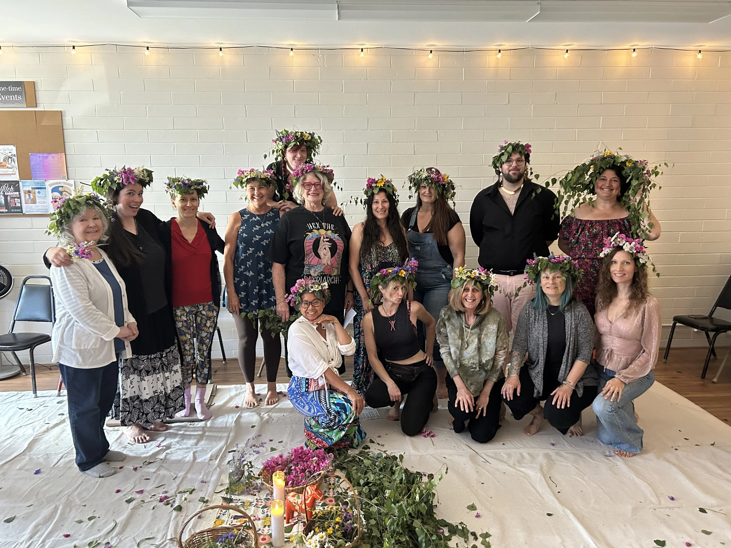 Group of people celebrating a floral event, wearing flower crowns, standing and kneeling on a white sheet with flowers, candles, and decorations in front of a white brick wall, with some chairs and a bulletin board visible.