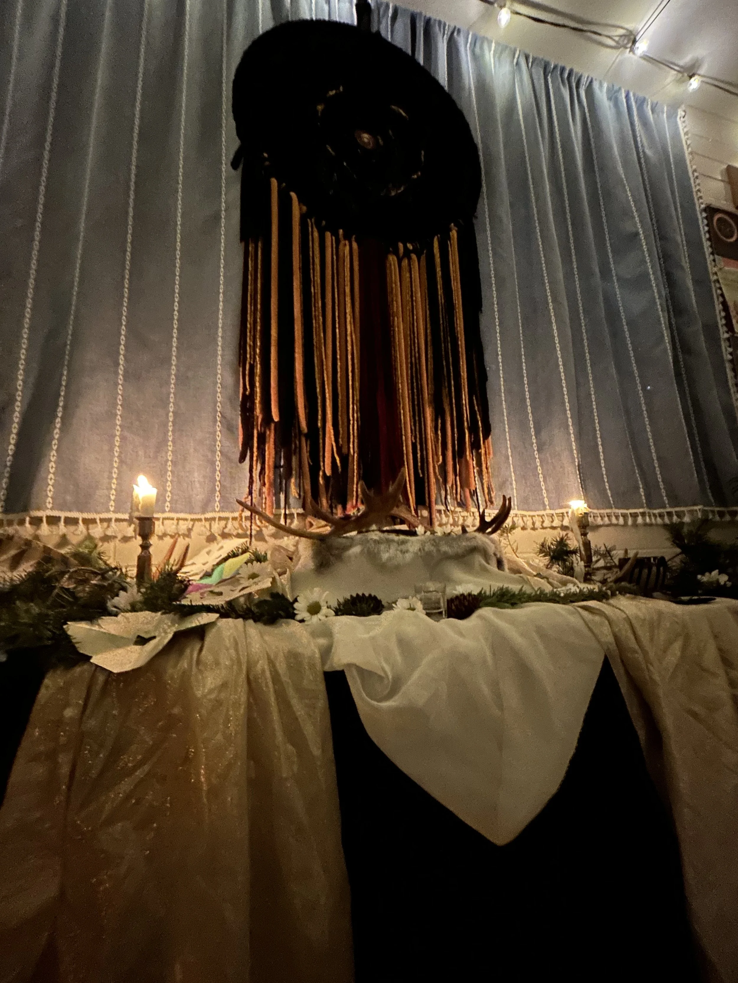 An indoor scene with a mounted deer head with antlers on a wall, beneath a large, suspended object resembling a rolled-up black blanket or fabric. The table is draped with white and gold cloths, decorated with flowers, candles, and greenery.