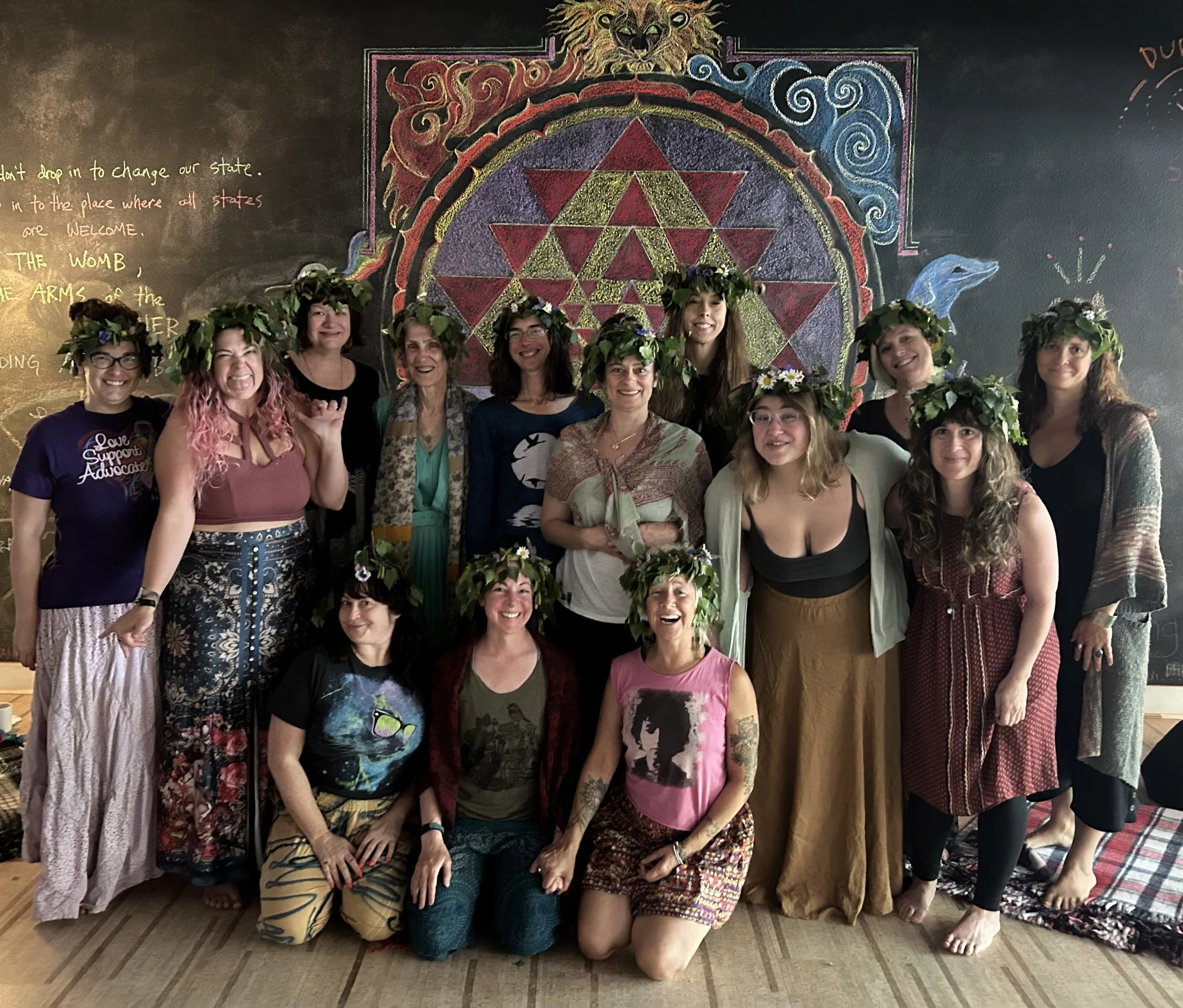Group of women wearing flower crowns, smiling, in front of a colorful chalkboard mural with geometric and spiritual symbols.