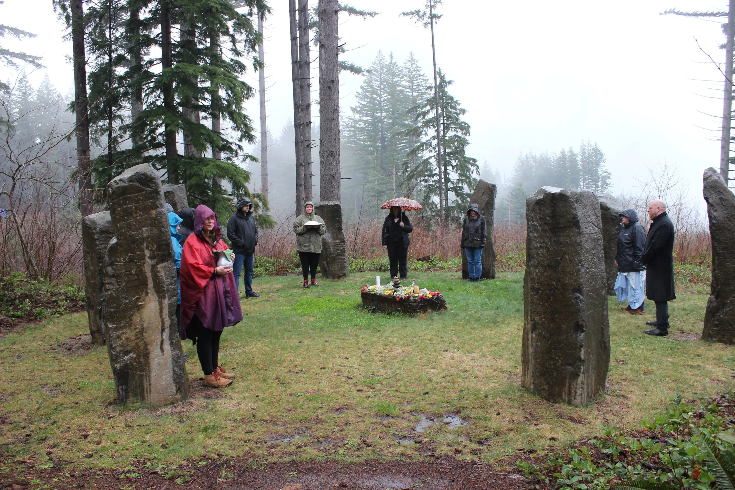 People gathered for a memorial service outdoors in a foggy forest, standing around a central altar with candles and flowers, surrounded by large standing stones.