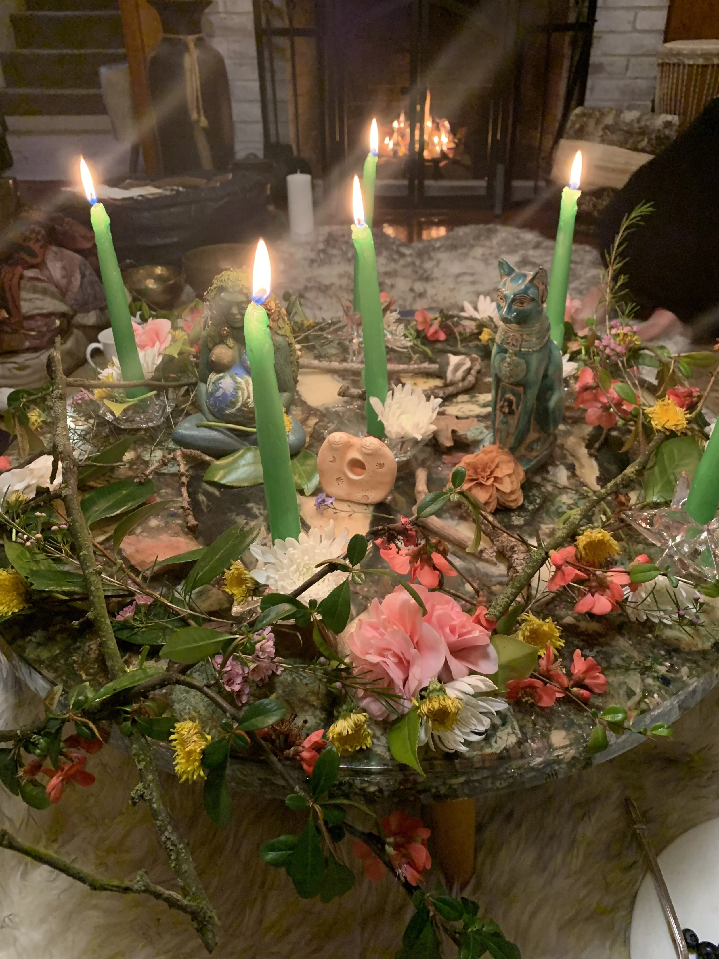 A table decorated with colorful flowers, green candles, and various decorative figurines with a lit fireplace in the background.