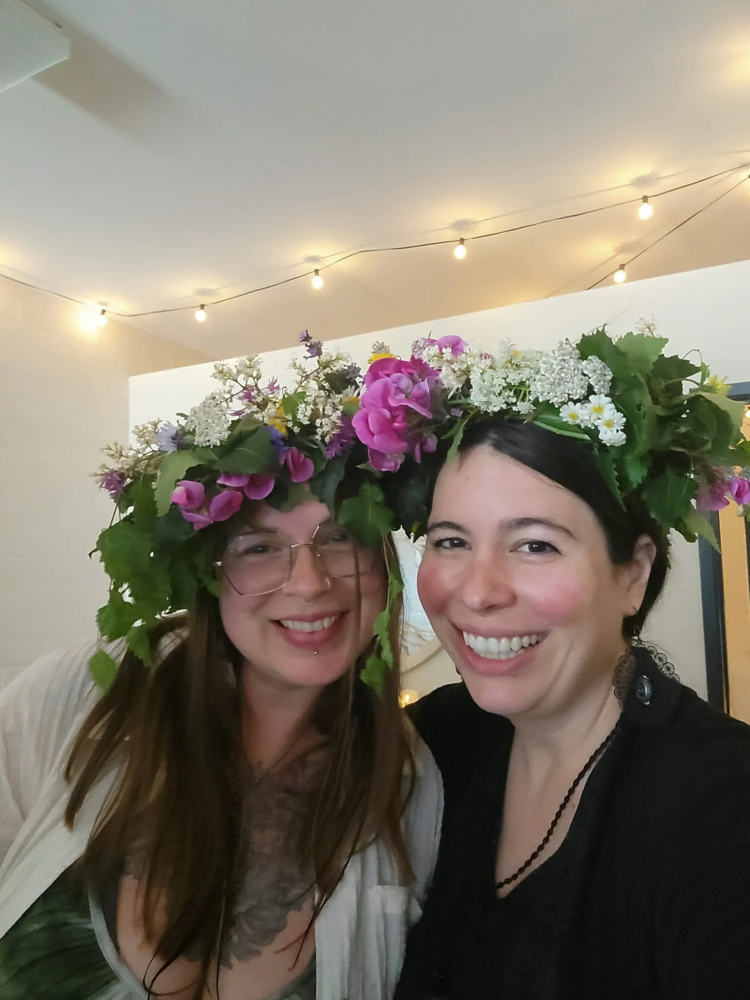 Two women smiling, wearing flower crowns made of various flowers and greenery, with string lights hanging in the background.