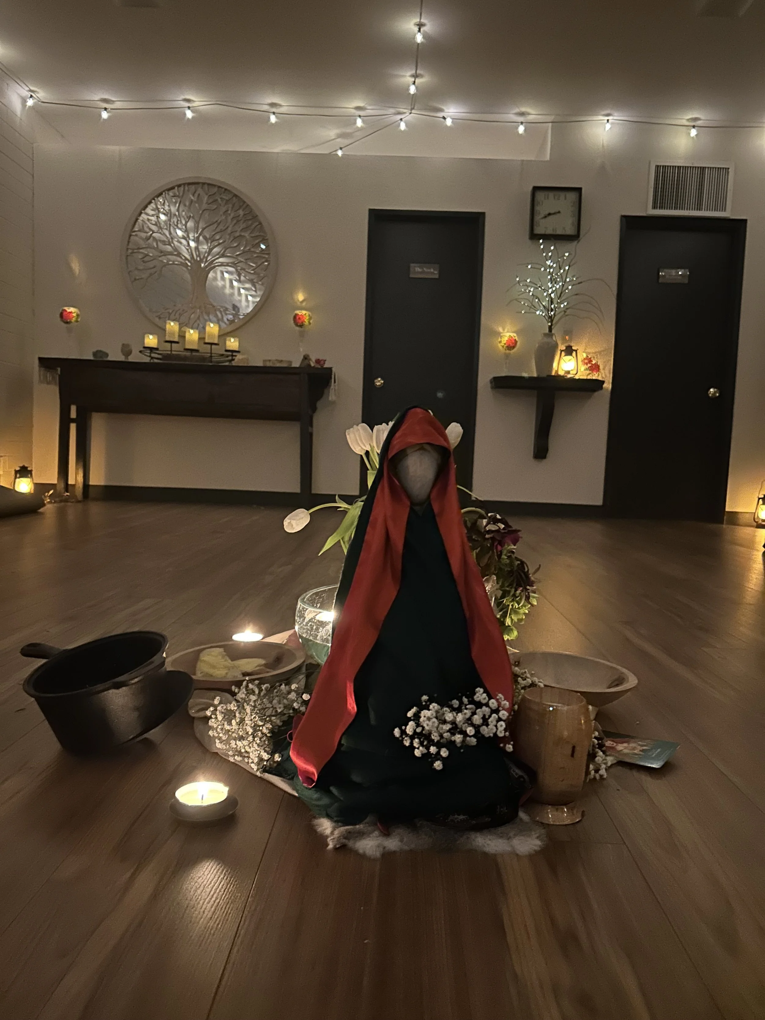 A decorative altar with a doll dressed in red and black, surrounded by flowers, candles, and bowls in a softly lit room with string lights and wall decor.