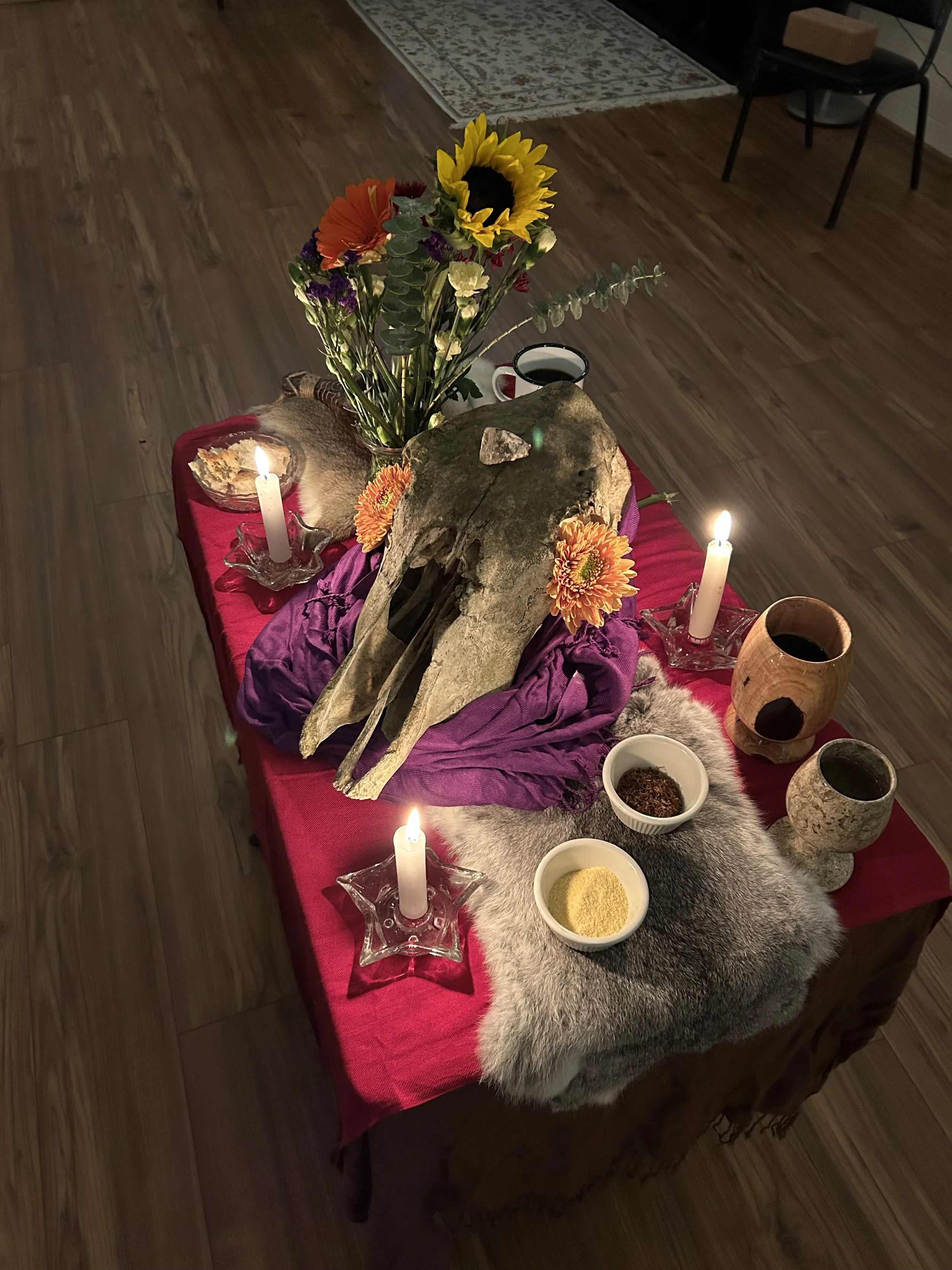 A table with a red cloth, candles, a skull, a bouquet of flowers, and various objects including bowls of powders and pottery, set on a fur and purple fabric, in a dimly lit room.