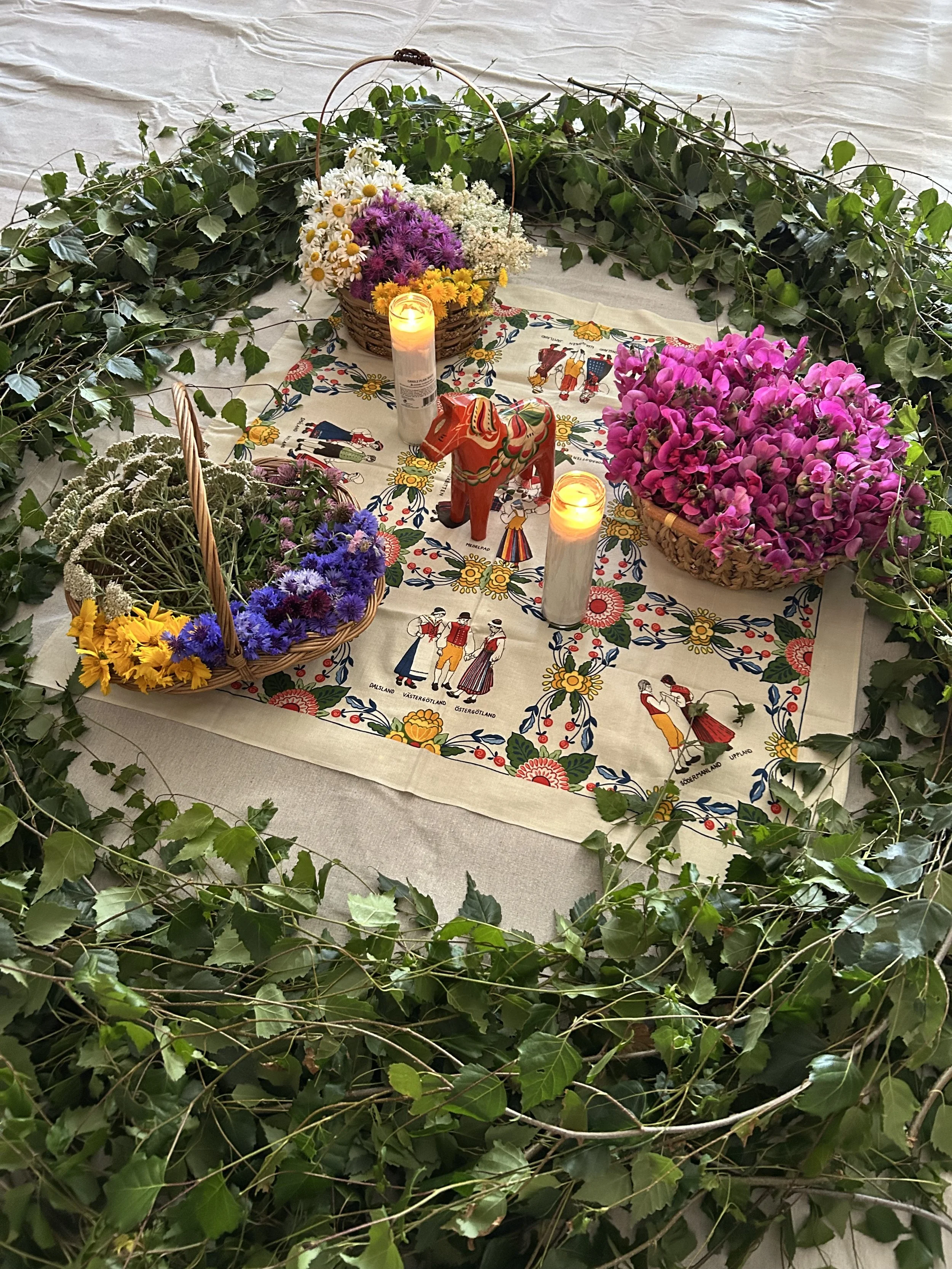 A table decorated with flowers, candles, and a colorful embroidered cloth, surrounded by greenery, with baskets of flowers and a decorative wooden horse in the center.