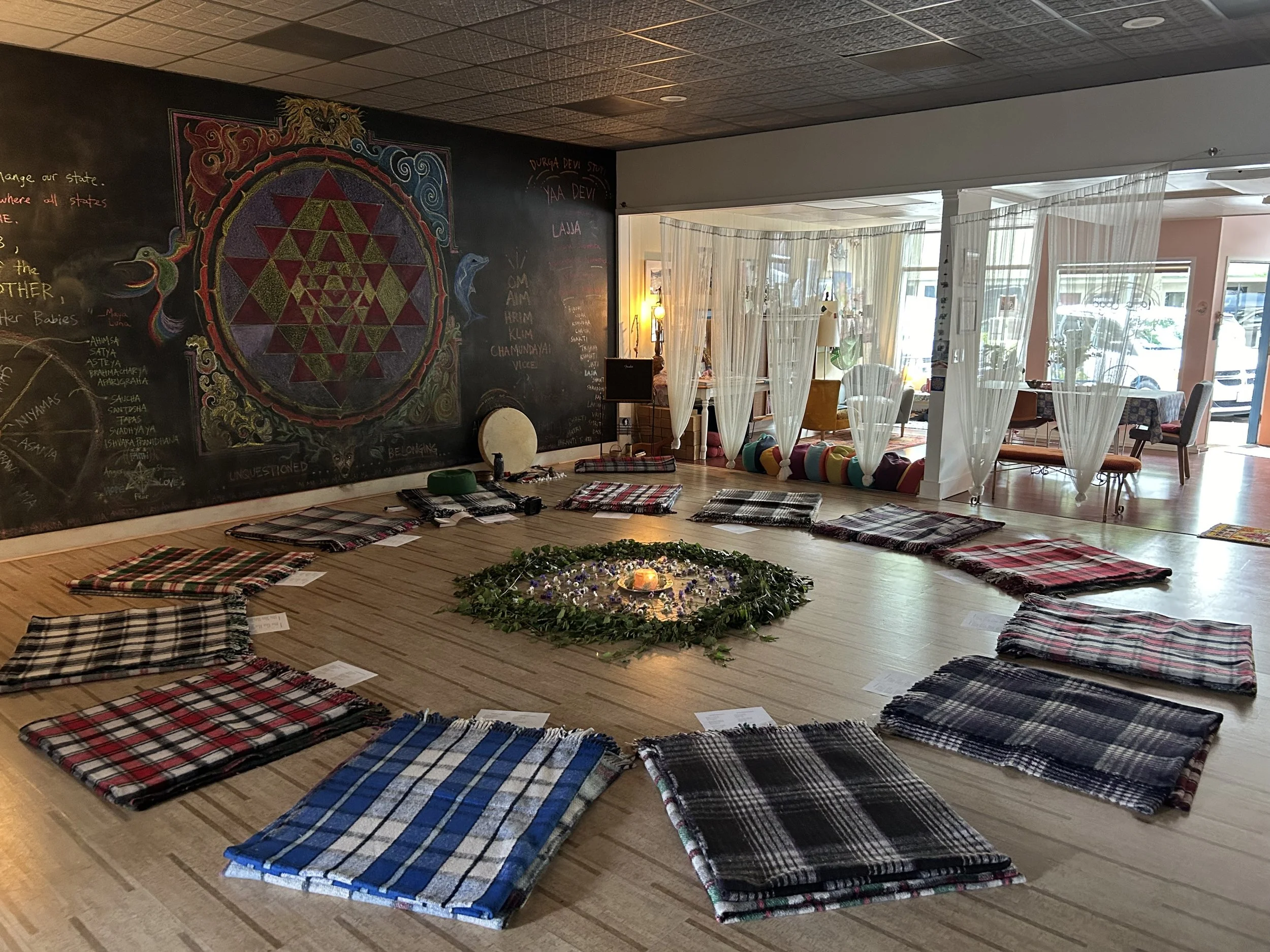 A circle of folded plaid blankets surrounds a small arrangement of greenery and candles on the floor of a cozy room, with curtains and chairs in the background.