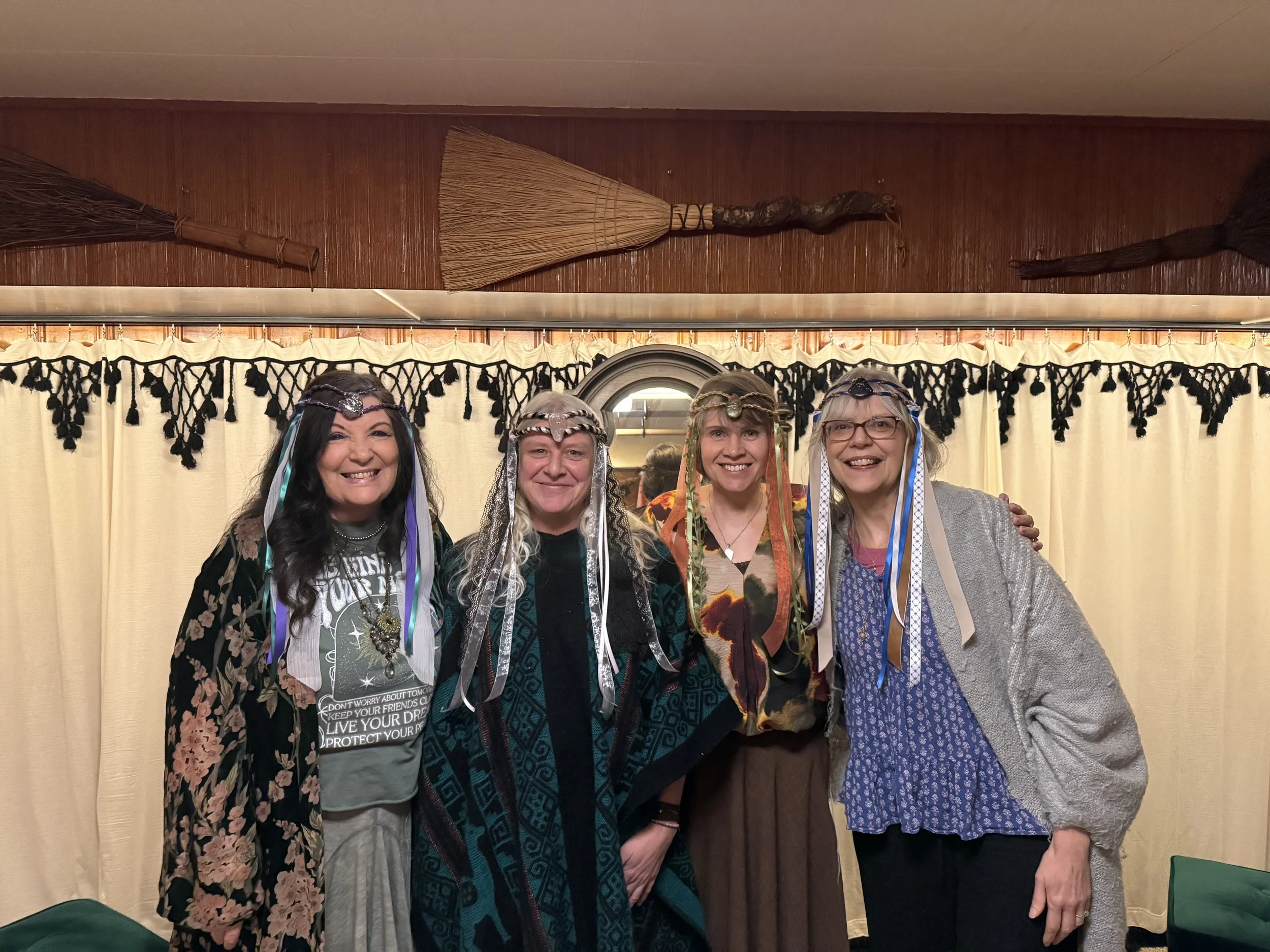 Four women dressed in bohemian or fairy-inspired costumes with crown-like headbands, standing together in front of a cream-colored curtain with black decorative trim, inside a room with wooden walls and broomstick decorations on the wall.