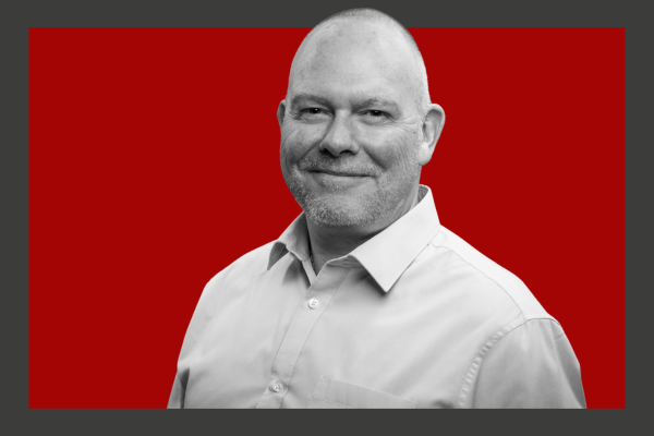 Scott Pawley: A middle-aged man with a bald head and beard, wearing a light-colored collared shirt, smiling slightly, against a red background.
