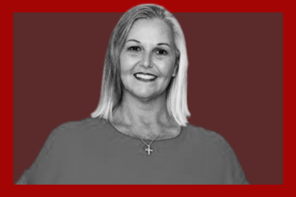 Andrea Caulfield-Smith: A woman with shoulder-length blonde hair, smiling, wearing a cross necklace and a top, against a red and dark background.